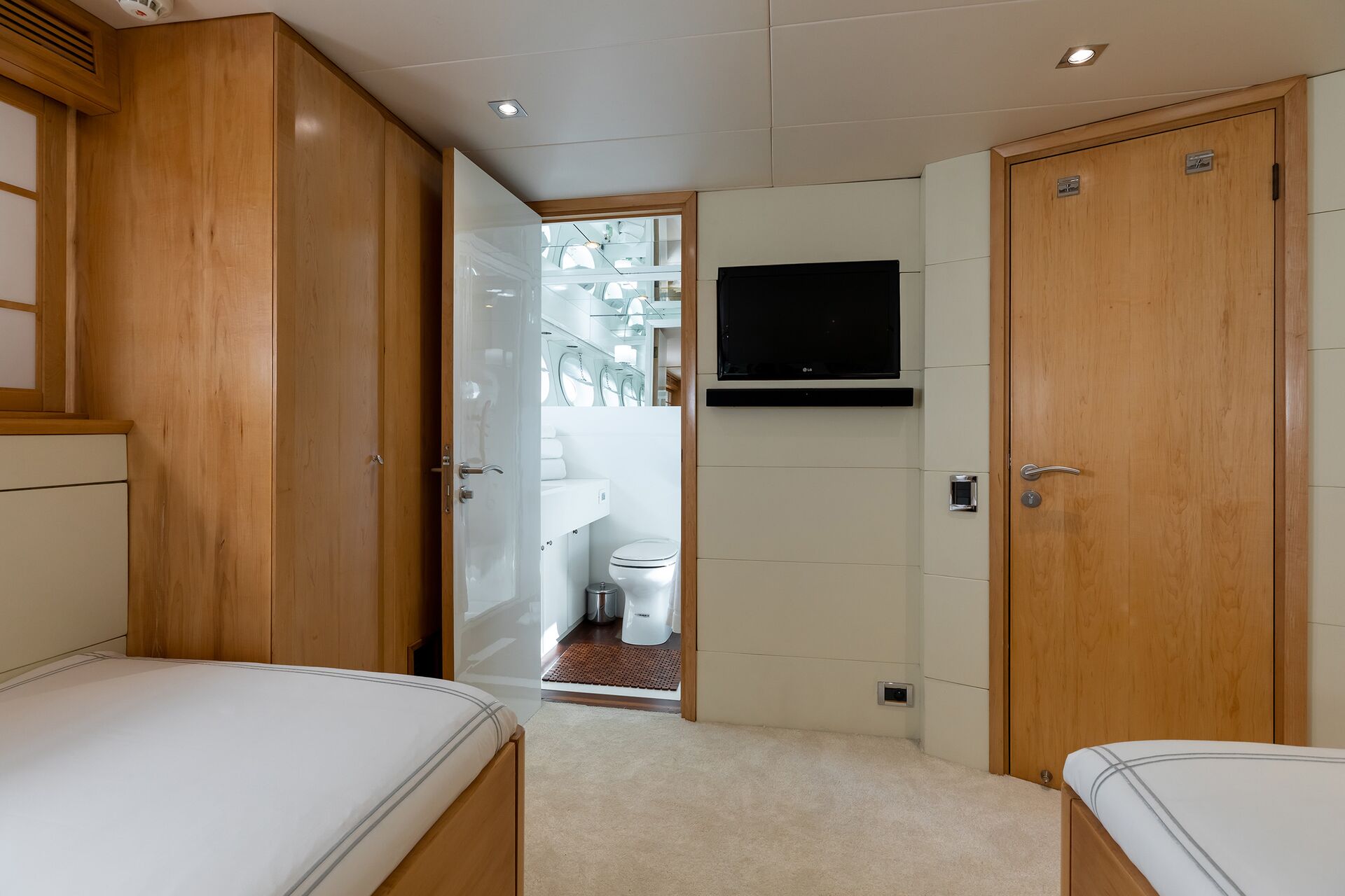 A modern, compact room with light wood paneling, featuring two single beds, a wall-mounted TV, and an open door leading to a white bathroom with a toilet and mirror.
