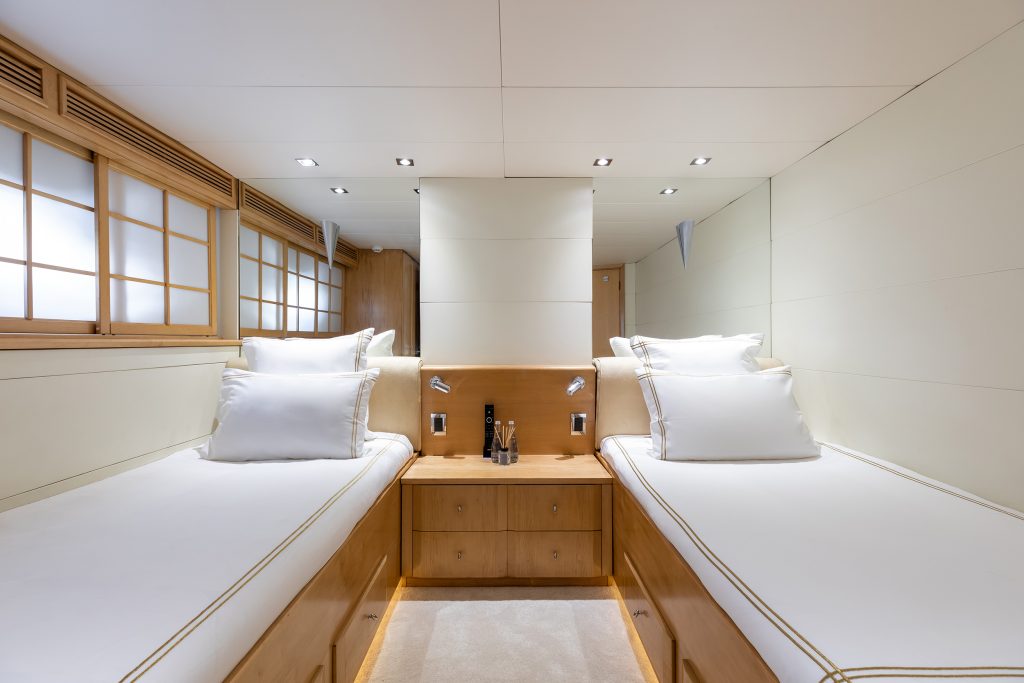 Modern yacht cabin with two single beds, white bedding, and wooden frames. A central nightstand with reading lights and water bottles separates the beds. Walls are light-colored, and there are wood-framed windows on one side.