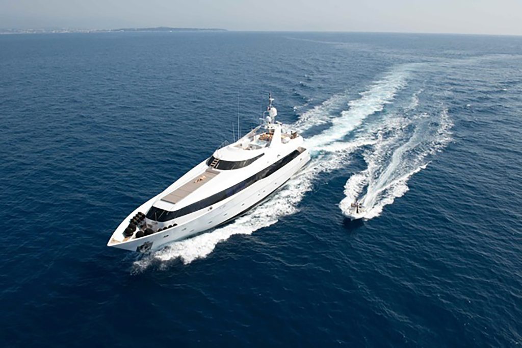 A large white luxury yacht cruises through open blue water, creating a wake, while a smaller motorboat trails alongside it under a clear sky.