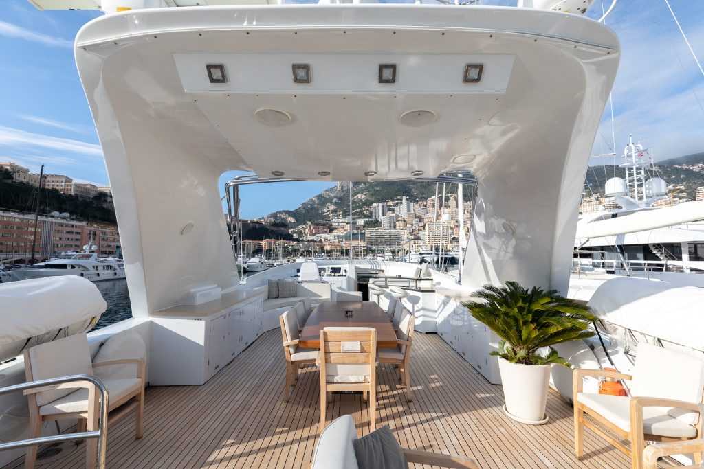 A spacious yacht deck with wooden flooring, outdoor dining table and chairs, potted plant, and harbor view with buildings and mountains in the background under a blue sky.