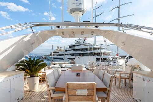 A luxurious yacht deck with a large wooden dining table, chairs, and potted plants, overlooking other yachts docked in a marina under a partly cloudy sky.