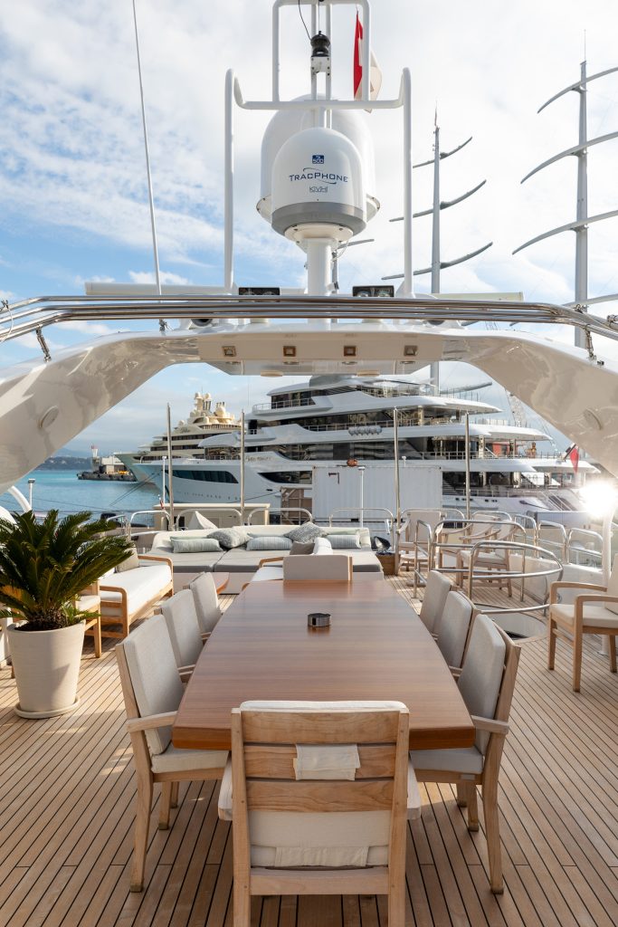A luxurious yacht deck with a wooden dining table and cushioned chairs, potted plants, and a view of other yachts and the ocean in the background under a partly cloudy sky.