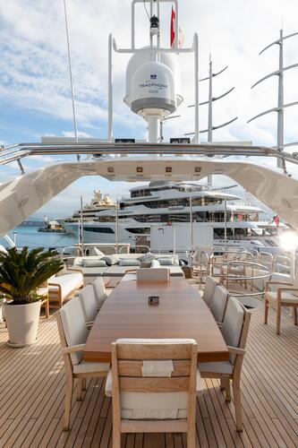 A luxurious yacht deck with a wooden dining table and cushioned chairs, potted plants, and a view of other yachts and the ocean in the background under a partly cloudy sky.