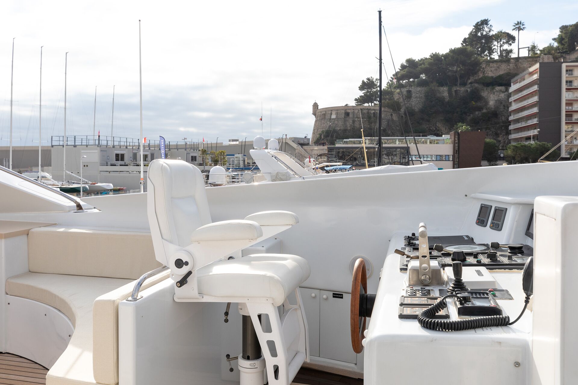 The image shows the control deck of a yacht, featuring a white captain’s chair, navigation controls, and a wooden steering wheel. In the background are buildings, trees, and docked boats at a marina under a partly cloudy sky.