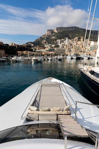 View from the bow of a luxury yacht docked in a marina, with several boats in the water and a city with tall buildings and hills in the background under a partly cloudy sky.
