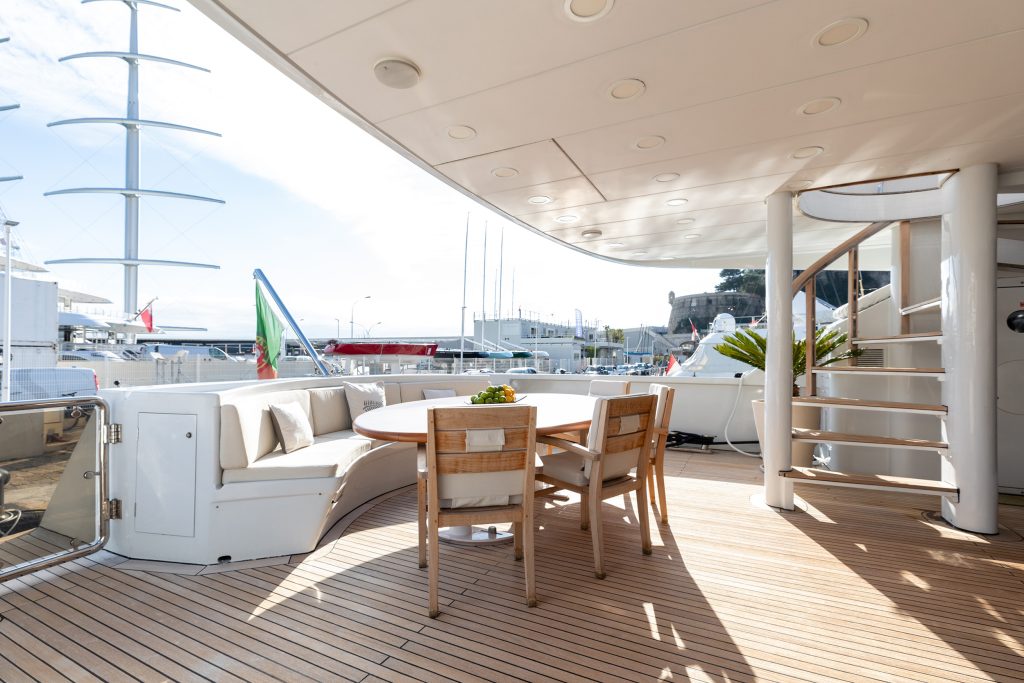 Outdoor yacht deck with wooden flooring, round table, chairs, curved white sofa, spiral staircase, and harbor view with boats and marina in the background under a clear sky.