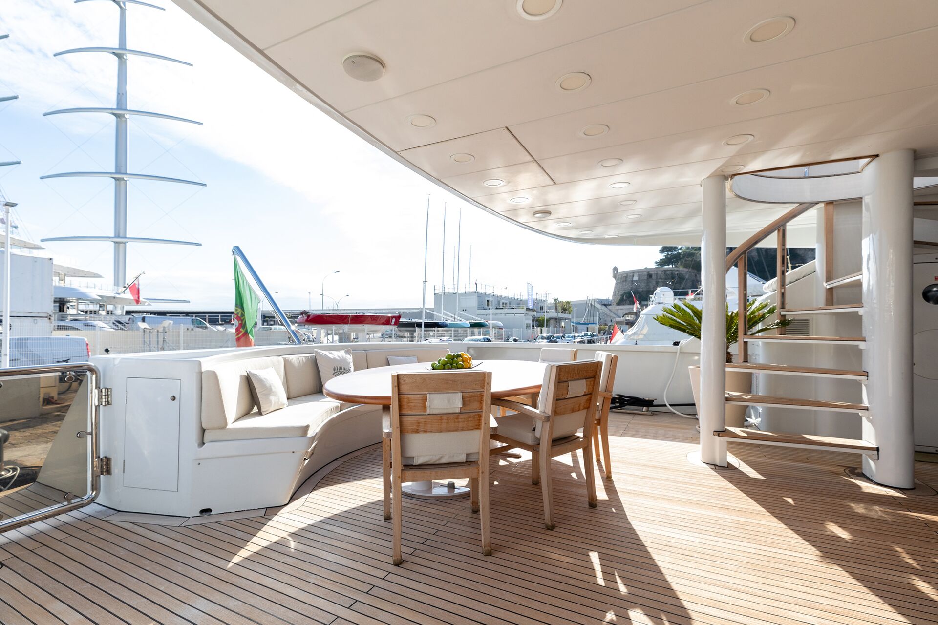 Outdoor yacht deck with wooden flooring, round table, chairs, curved white sofa, spiral staircase, and harbor view with boats and marina in the background under a clear sky.
