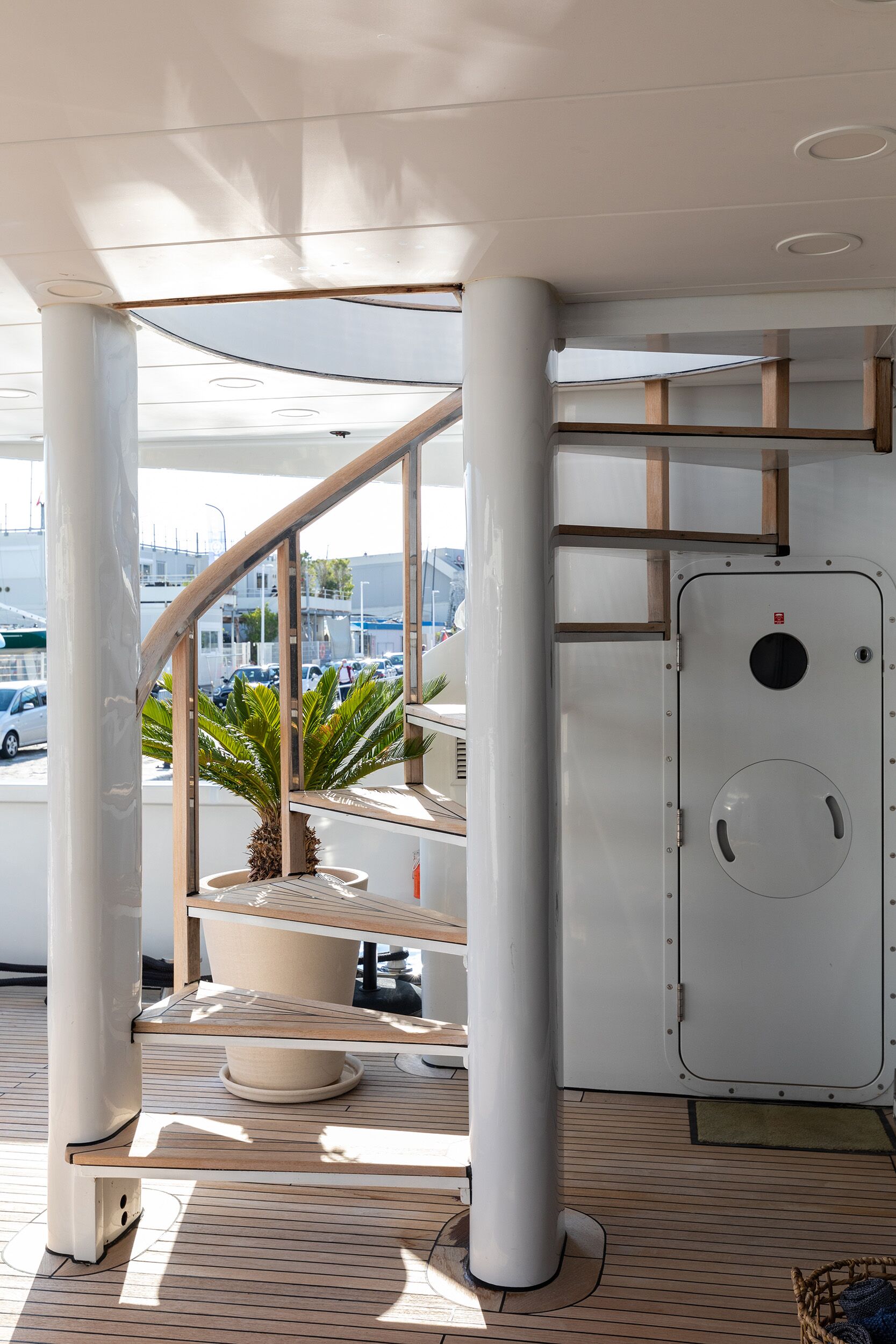 A modern yacht deck with a spiral wooden staircase beside a white door. A potted palm sits near the stairs, and sunlight streams onto the wooden floor. Marina and parked cars are visible outside.