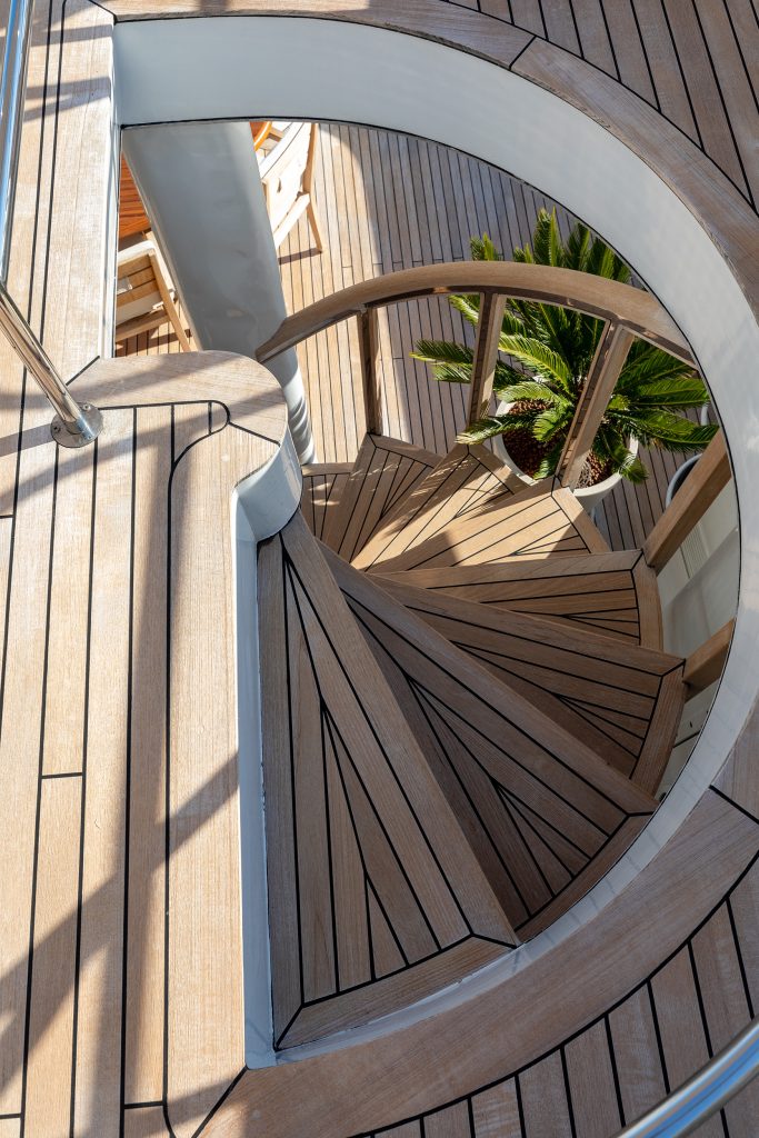 Curved wooden staircase with black trim, leading down to a lower deck. A potted palm plant is visible at the bottom of the stairs, and the surrounding area features matching wooden panel flooring.