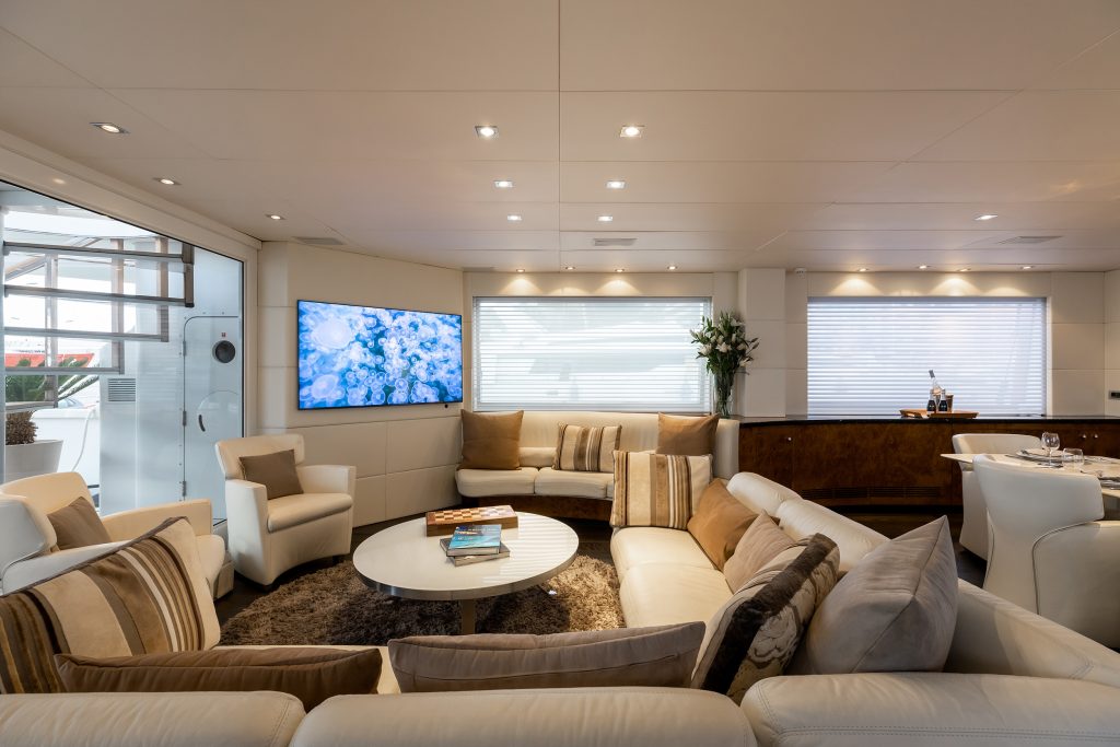 A luxurious yacht lounge with modern white and beige furnishings, a round coffee table, armchairs, a TV displaying underwater scenes, large windows with blinds, and a bar area with drinks and flowers.