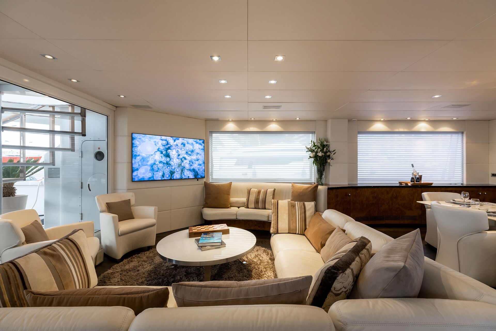 A luxurious yacht lounge with modern white and beige furnishings, a round coffee table, armchairs, a TV displaying underwater scenes, large windows with blinds, and a bar area with drinks and flowers.