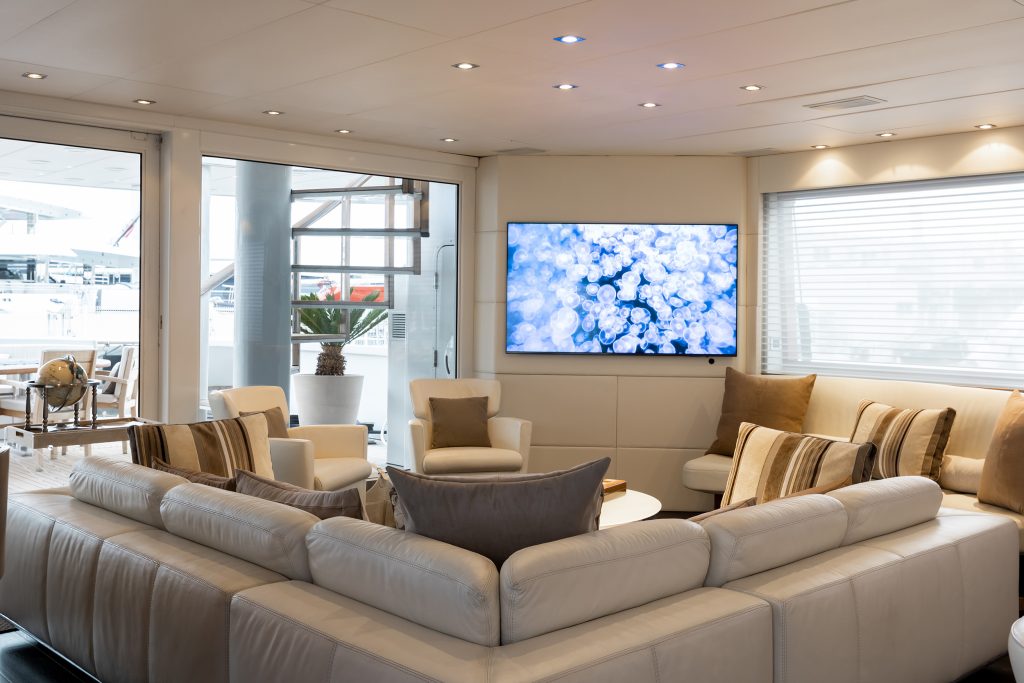 Modern living room with a large white sectional sofa, beige and brown cushions, a wall-mounted TV displaying an abstract blue image, armchairs, and large windows letting in natural light.
