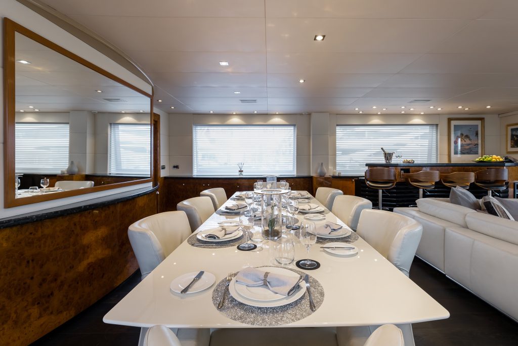 A modern dining area with a glossy white table set for eight, surrounded by white chairs. Behind the table is a large mirror, and in the background, a lounge with bar stools and a sofa, all in a well-lit, elegant room.