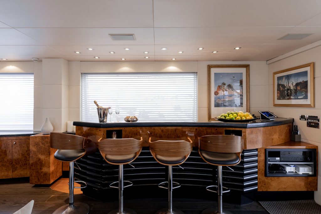 Modern home bar with four wooden stools, a curved counter, a bowl of green apples, champagne in an ice bucket, framed art on the walls, and a window with closed blinds in a sleek, well-lit room.