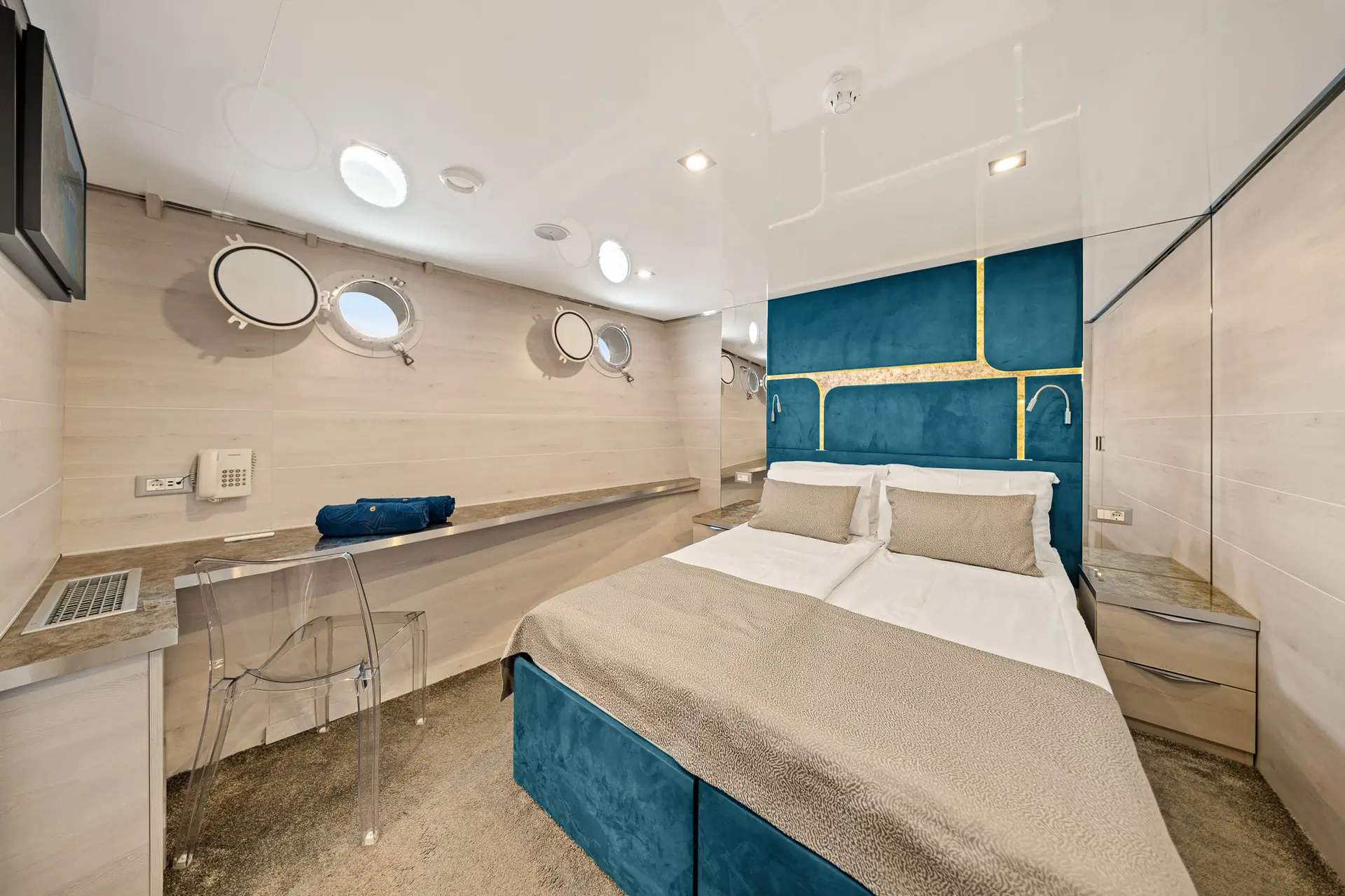 A modern yacht cabin with a double bed, blue and beige decor, round porthole windows, a clear acrylic chair, wall-mounted TV, and folded towels on a long desk.