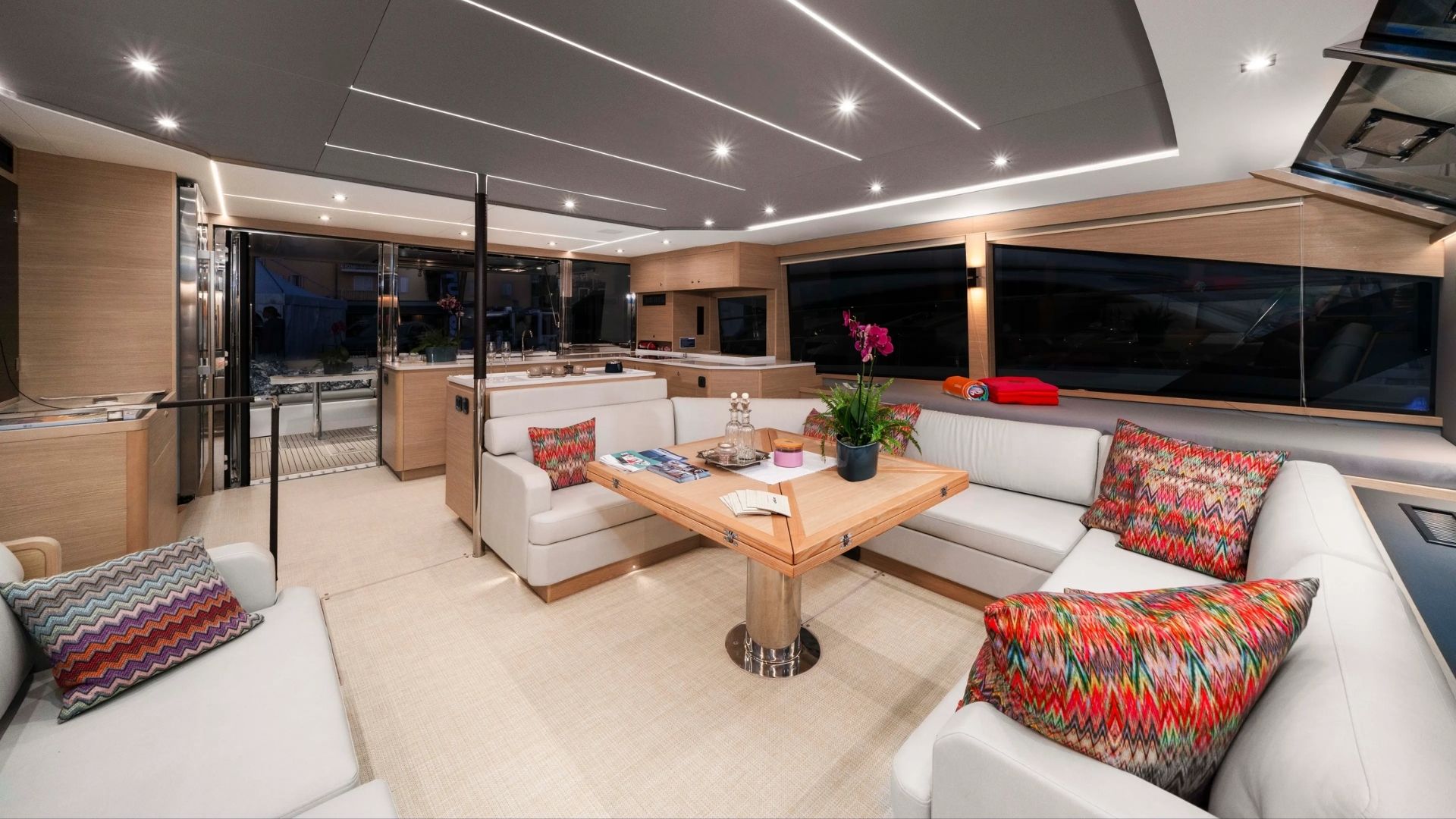 Modern yacht interior featuring light wood finishes, a central wooden dining table, white sofas with colorful patterned pillows, built-in kitchen appliances, potted plants, and large windows with nighttime views.