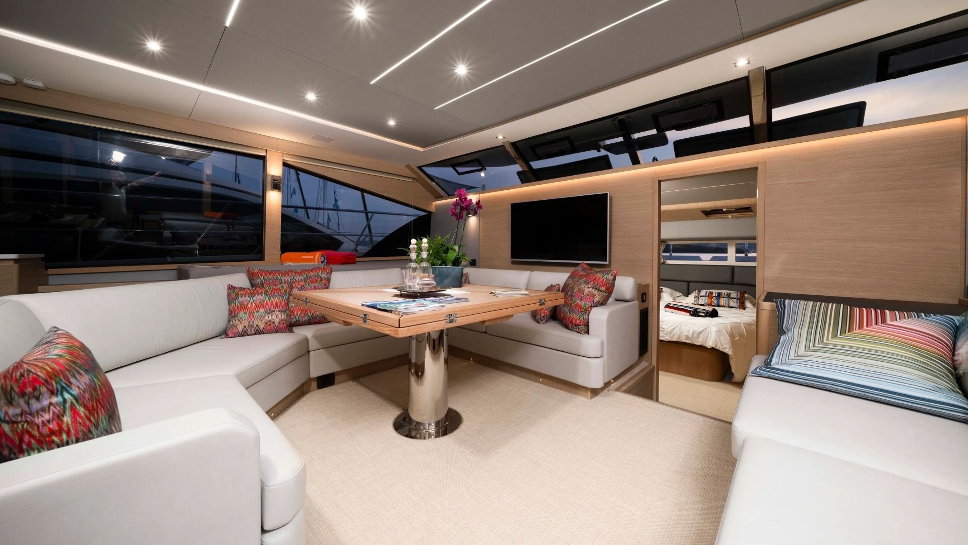 Luxurious yacht interior with light wood finishes, white sofas, colorful pillows, a central table with plants, large windows, and a doorway leading to a cozy bedroom with a neatly made bed.