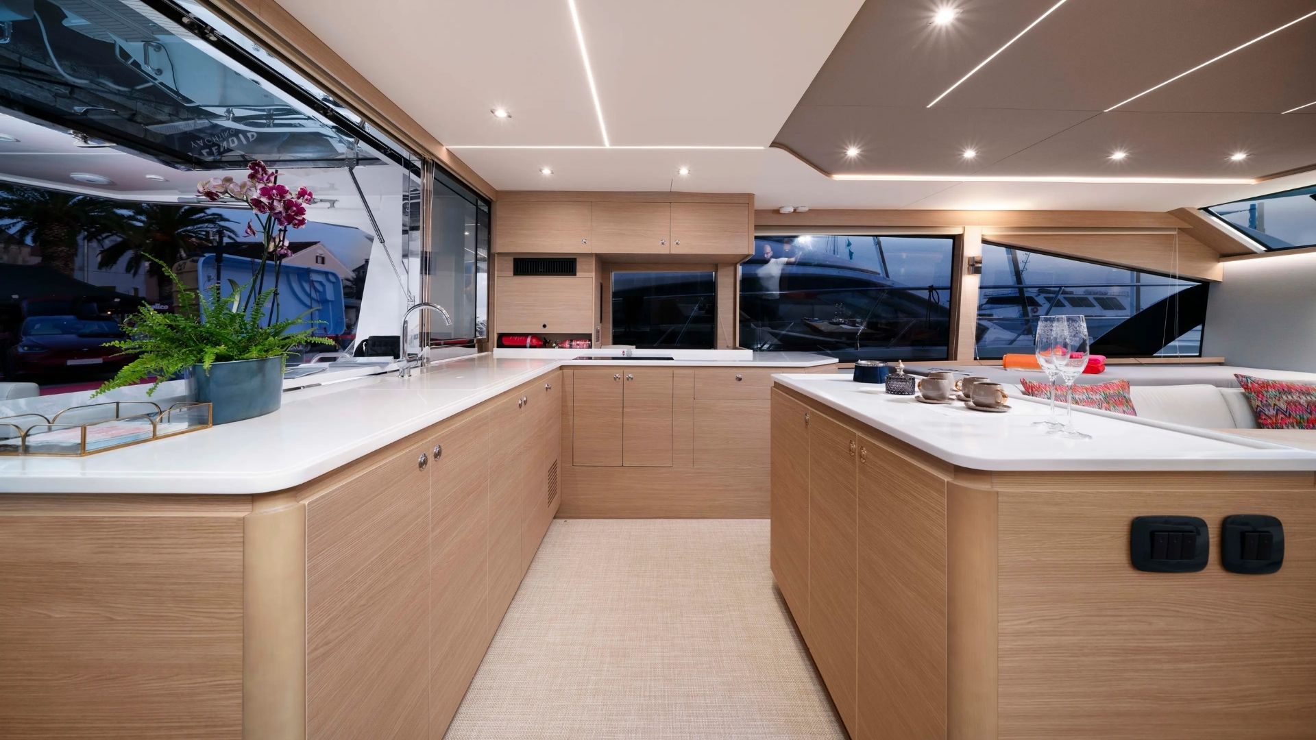 Modern yacht interior with light wood cabinetry, white countertops, large windows, and decorative plants. There are wine glasses and tableware set on the counter, with cushioned seating visible on the right.