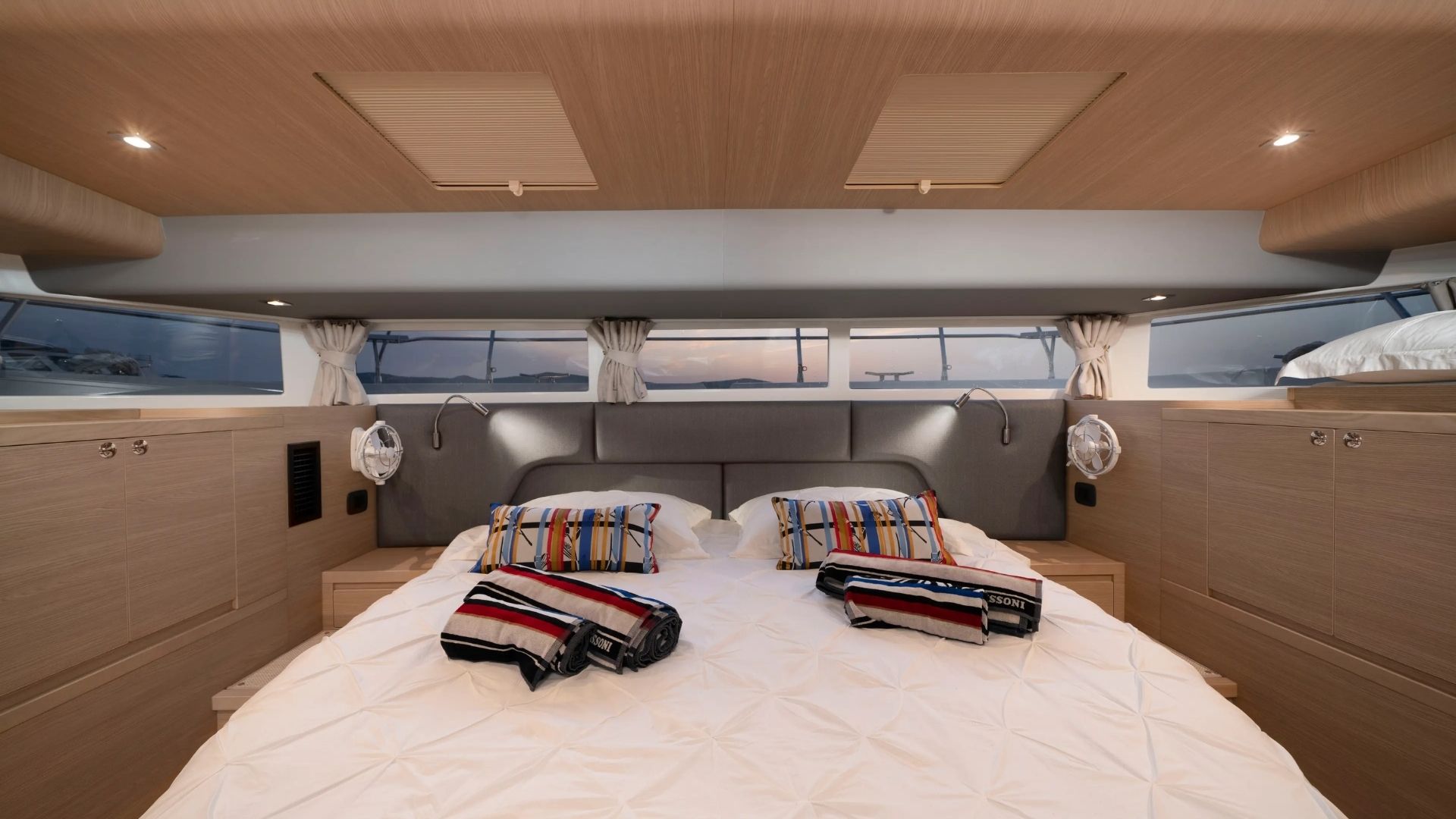 A cozy bedroom on a yacht with a large bed, striped pillows, and folded towels. There are large windows with curtains, built-in lights, and wooden cabinetry around the room.