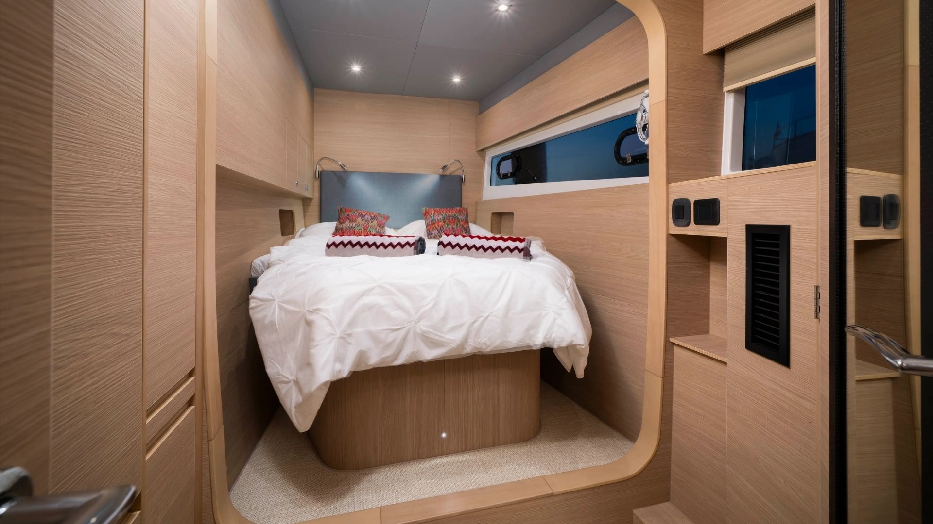A modern, compact bedroom with light wood walls and ceiling, a neatly made double bed with white bedding and patterned pillows, wall-mounted reading lights, and a large window beside the bed.
