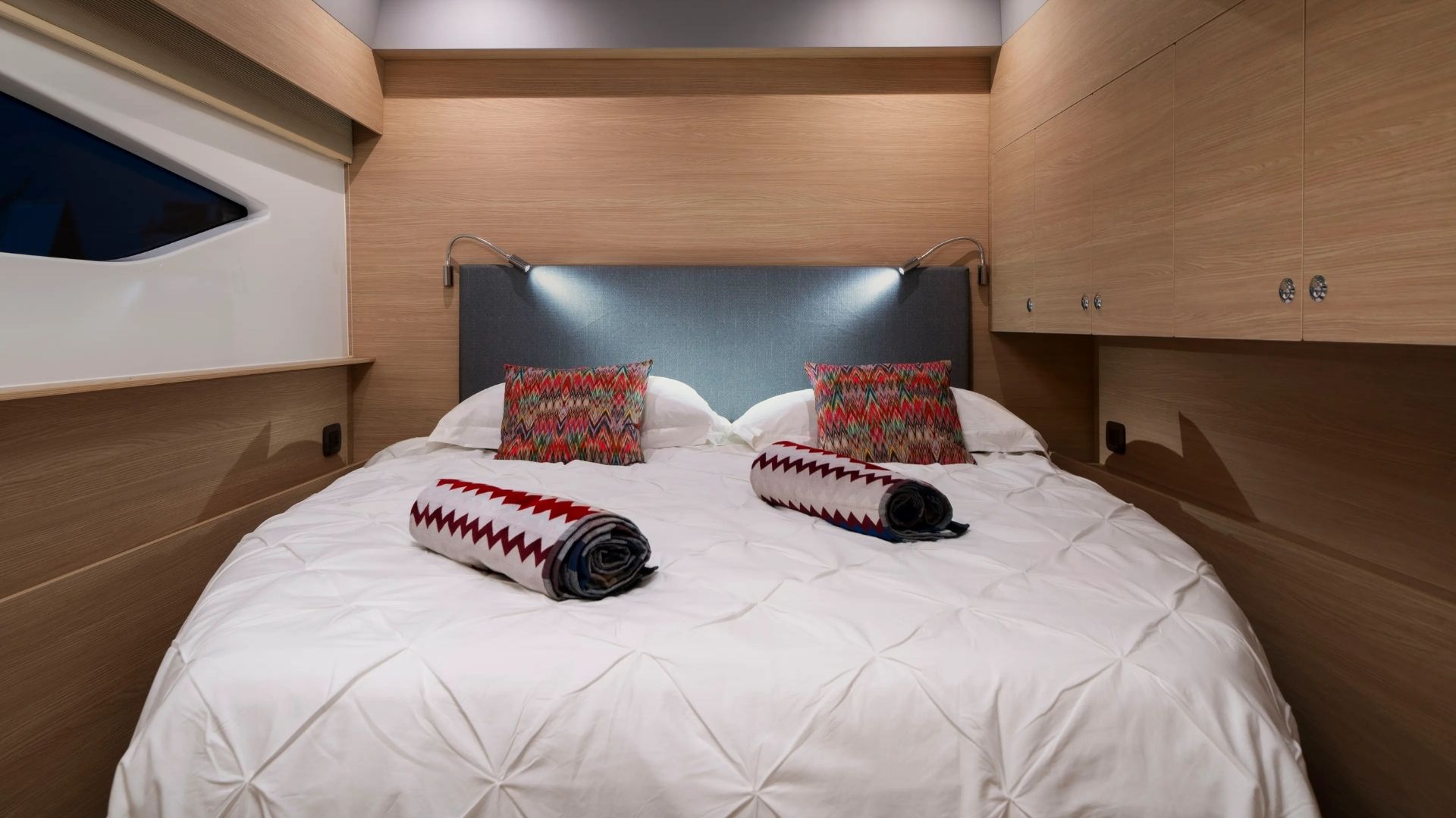 A cozy bedroom with a large bed, white comforter, two patterned pillows, and two rolled blankets. Built-in wooden cabinets line the walls and modern reading lights are mounted above the headboard.