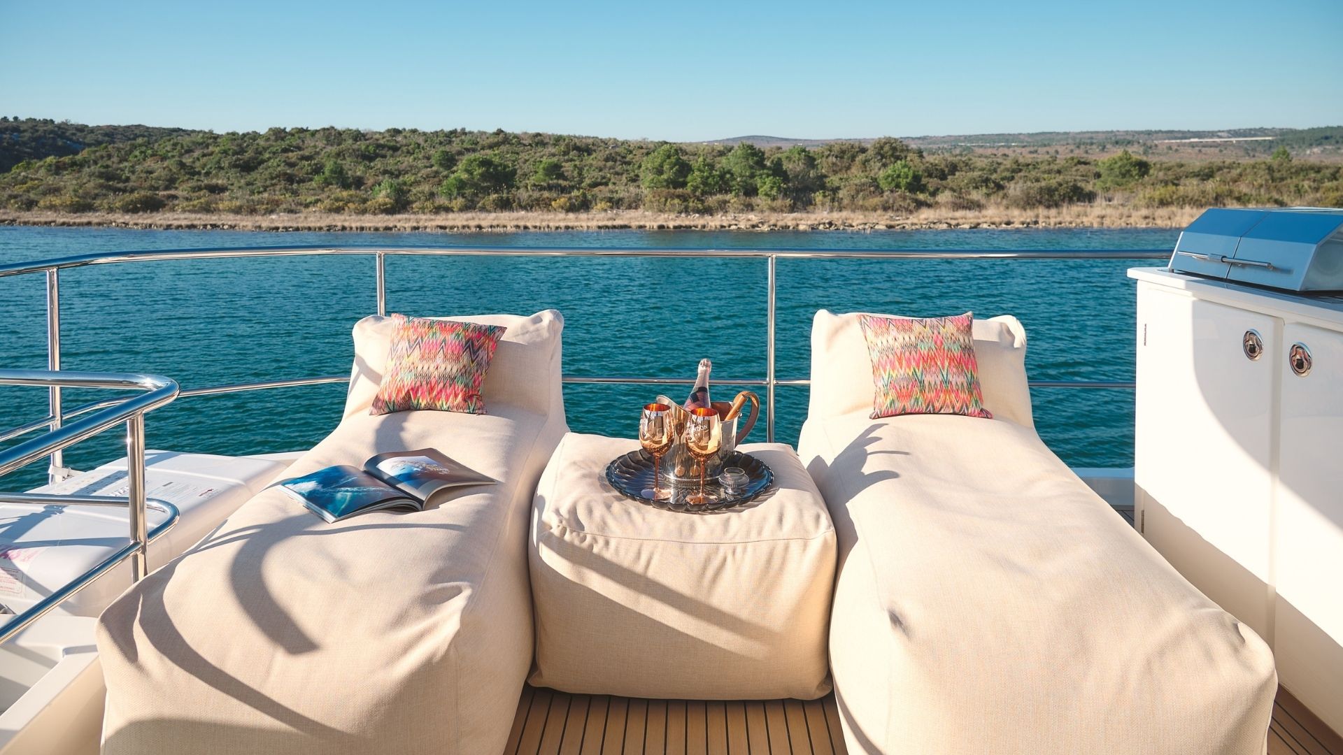 Two beige lounge chairs with colorful pillows on a yacht deck face calm blue water and a green shoreline. A round table between them holds a silver tray with drinks. A magazine and grill are also visible.