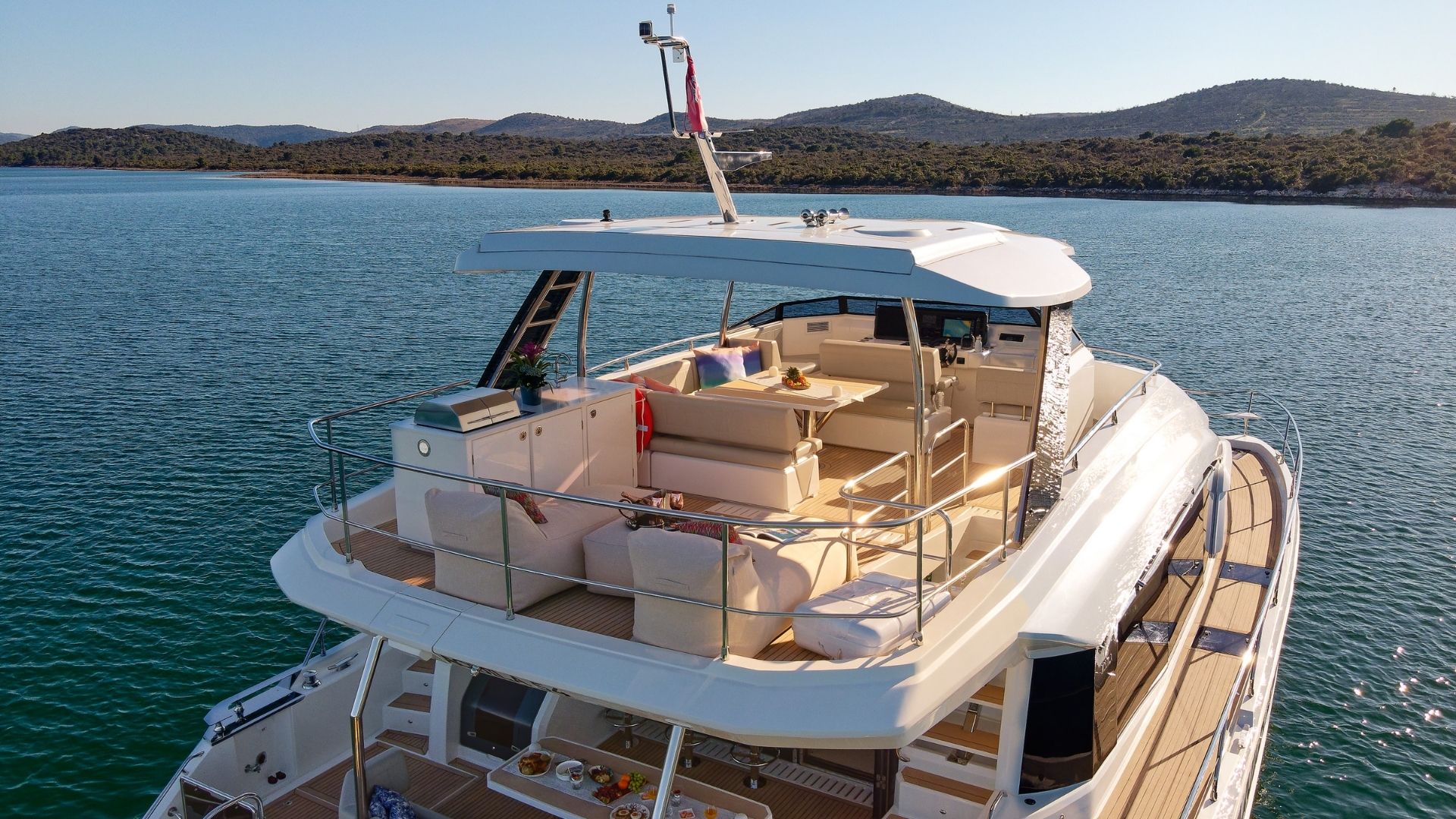 A luxurious yacht anchored on calm water, featuring an upper deck with cushioned seating, a table, and open views of the surrounding hilly shoreline under a clear sky.