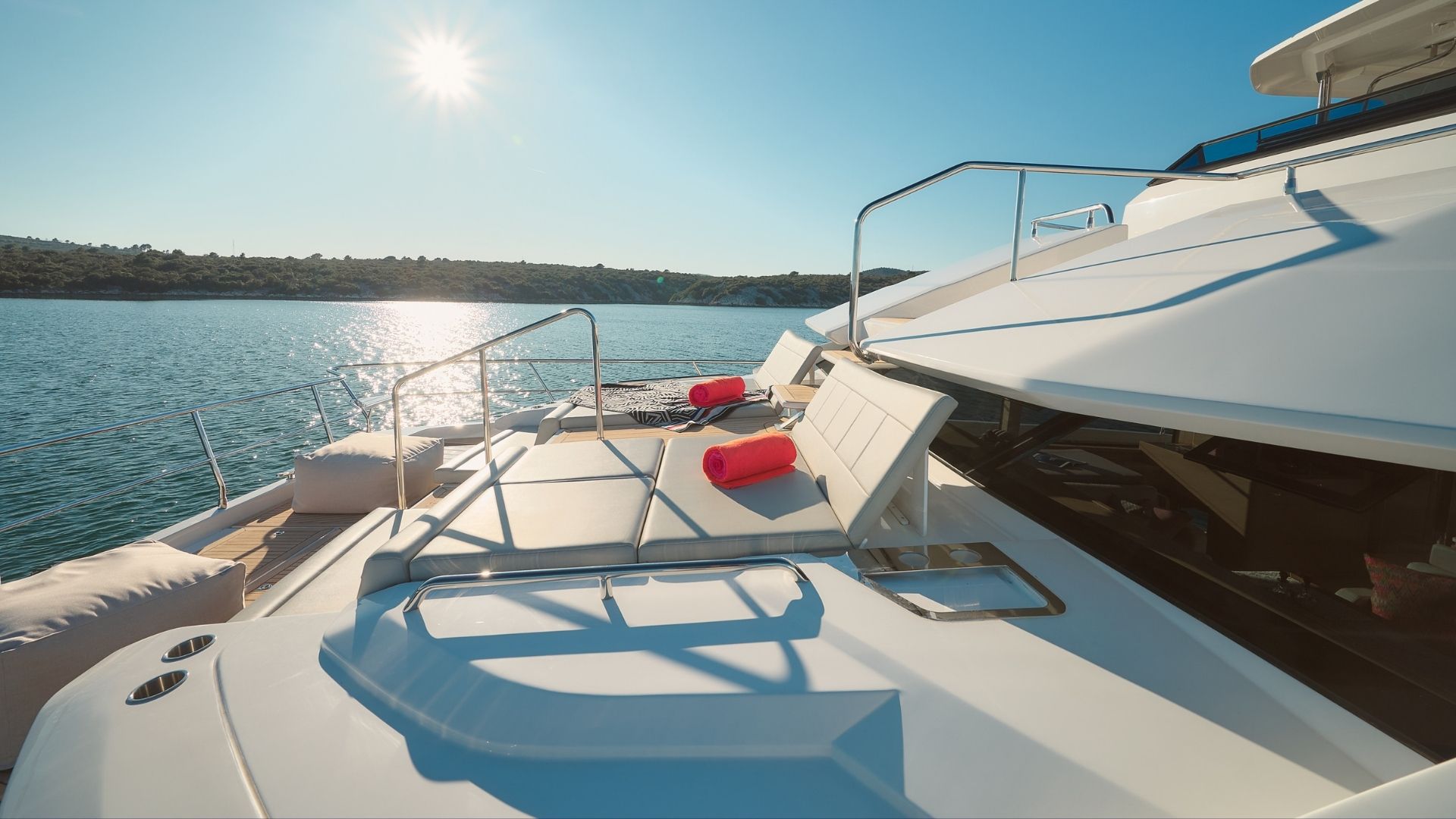 A luxurious yacht deck with white lounge chairs and red towels overlooks calm blue water under a sunny, clear sky. Land with green vegetation is visible in the background.