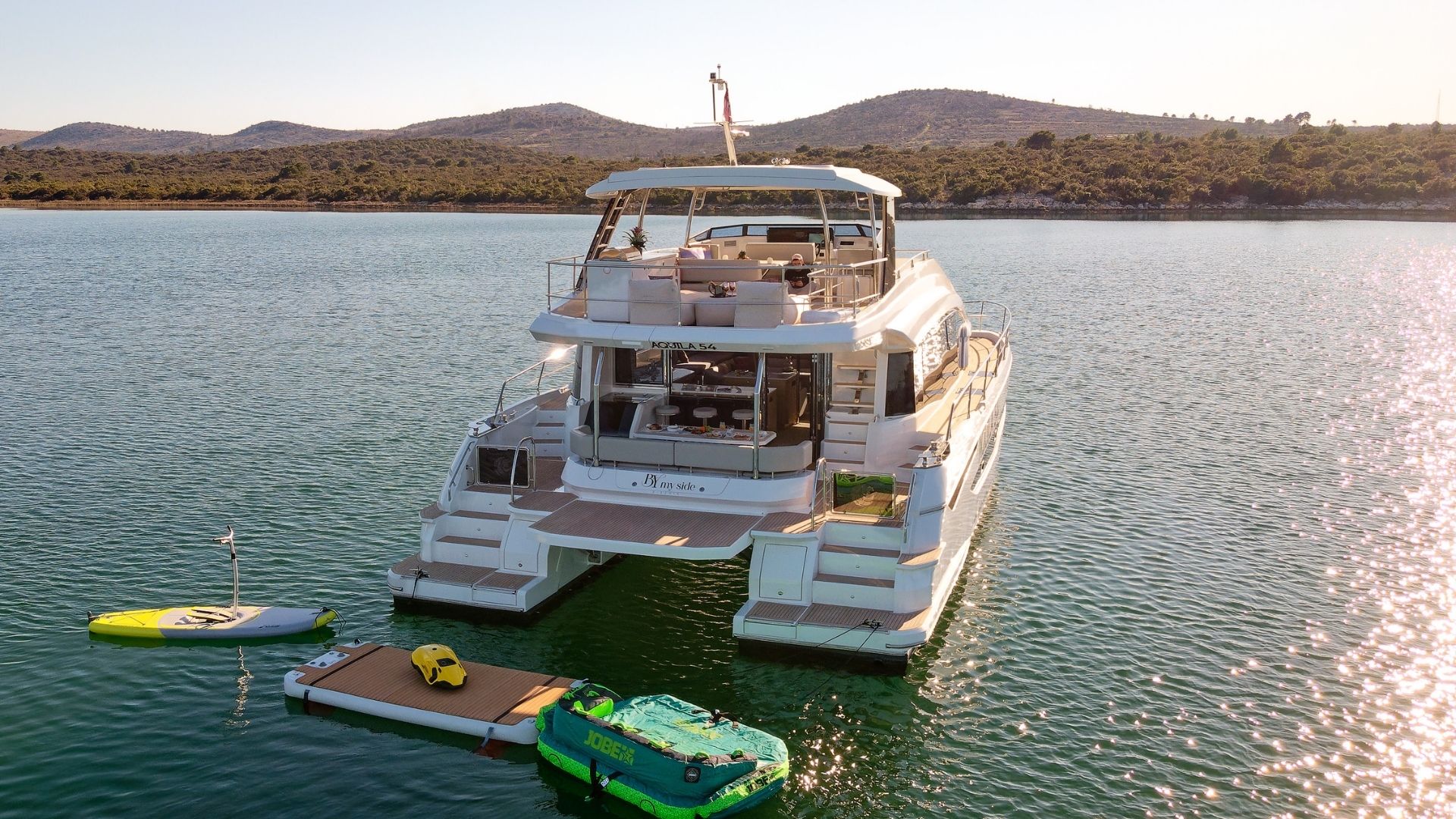 A luxury white yacht is anchored on calm water near a tree-lined shore with hills in the background. Inflatable water toys, a paddleboard, and a floating dock are beside the yacht. The sun is low in the sky.