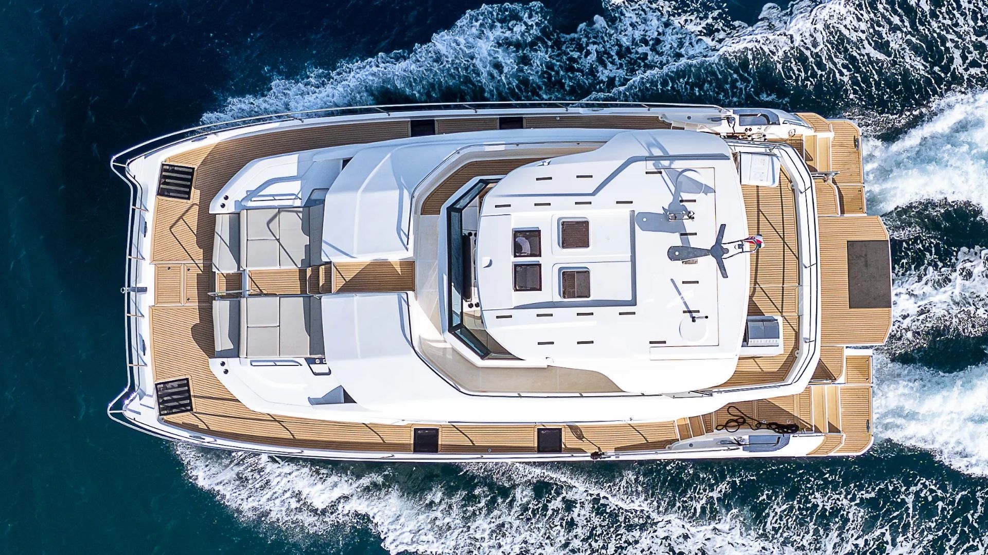 Aerial view of a modern white yacht with wooden decking cruising through deep blue water, leaving a trail of white waves behind.