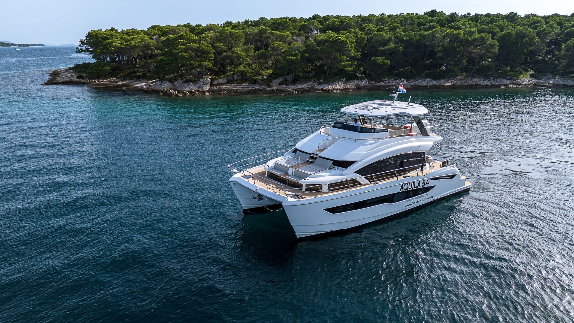 A white luxury yacht is anchored in calm blue water near a small, forested island with rocky shores and dense green trees under a clear sky.