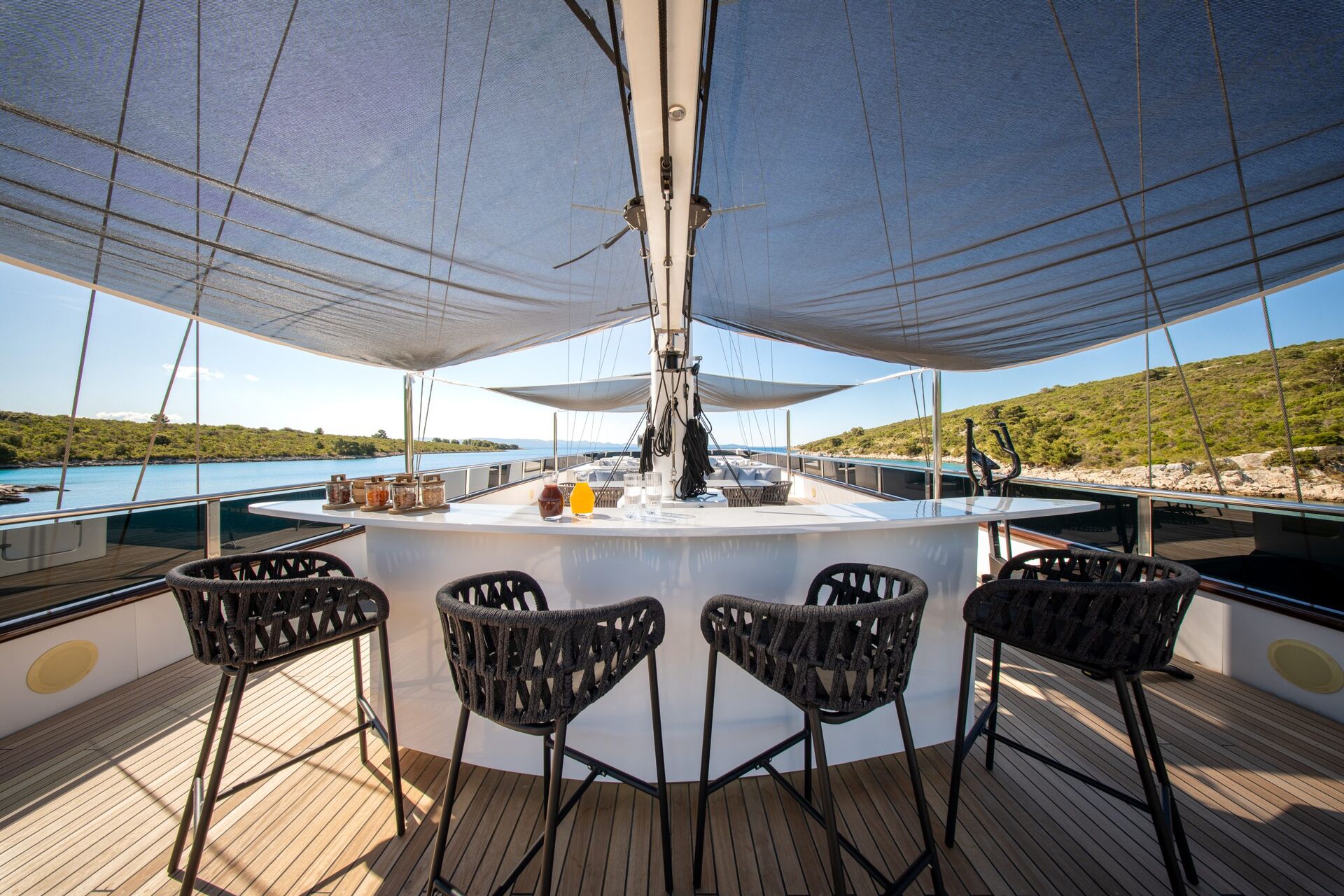 A stylish outdoor bar with four woven bar stools on a yacht deck, shaded by blue canopies, overlooks calm water and green hills under a clear sky.