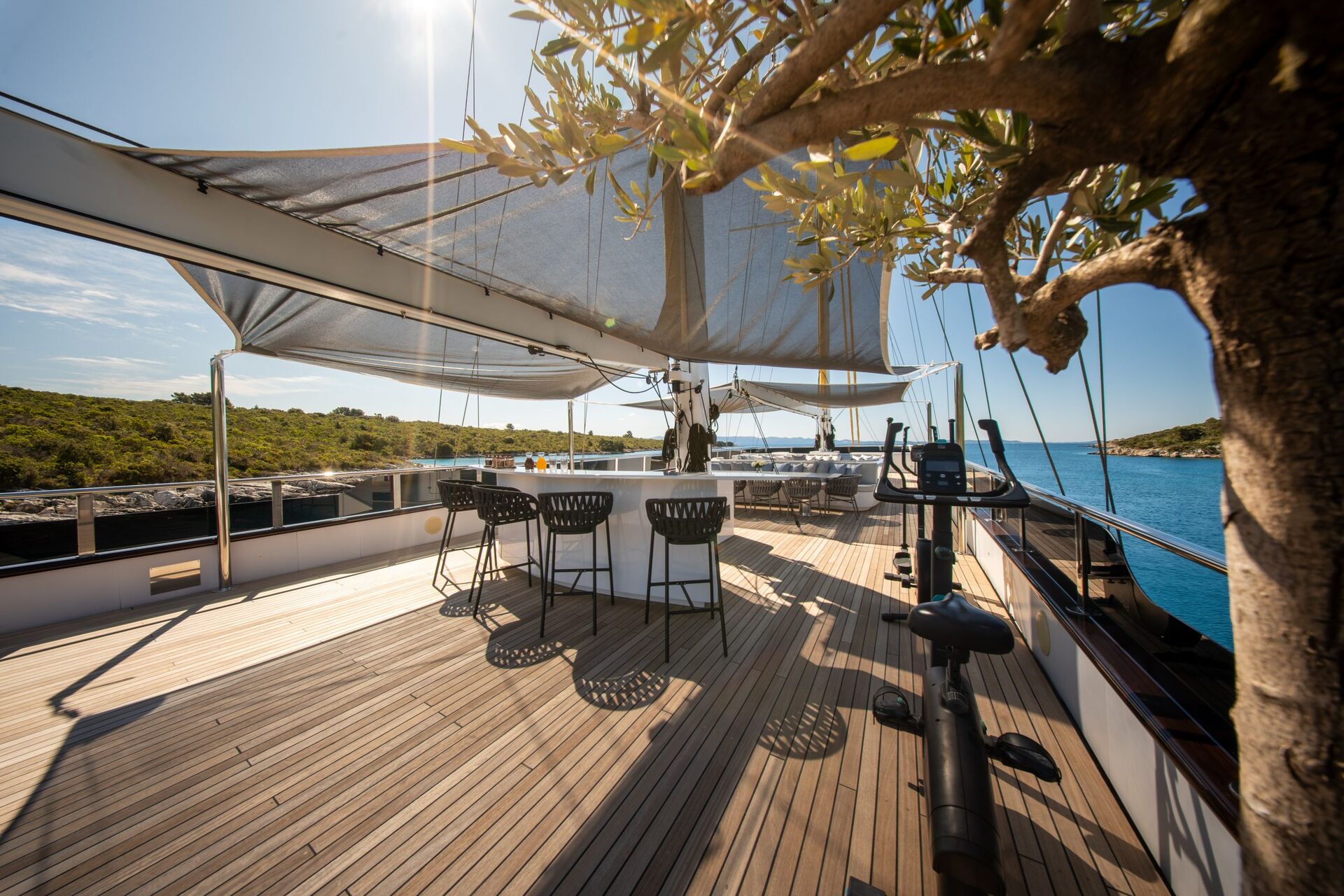 Sunny deck of a yacht with wooden flooring, a shaded seating area with chairs around a bar table, an exercise bike, and a tree in the foreground, overlooking blue water and a green coastline.