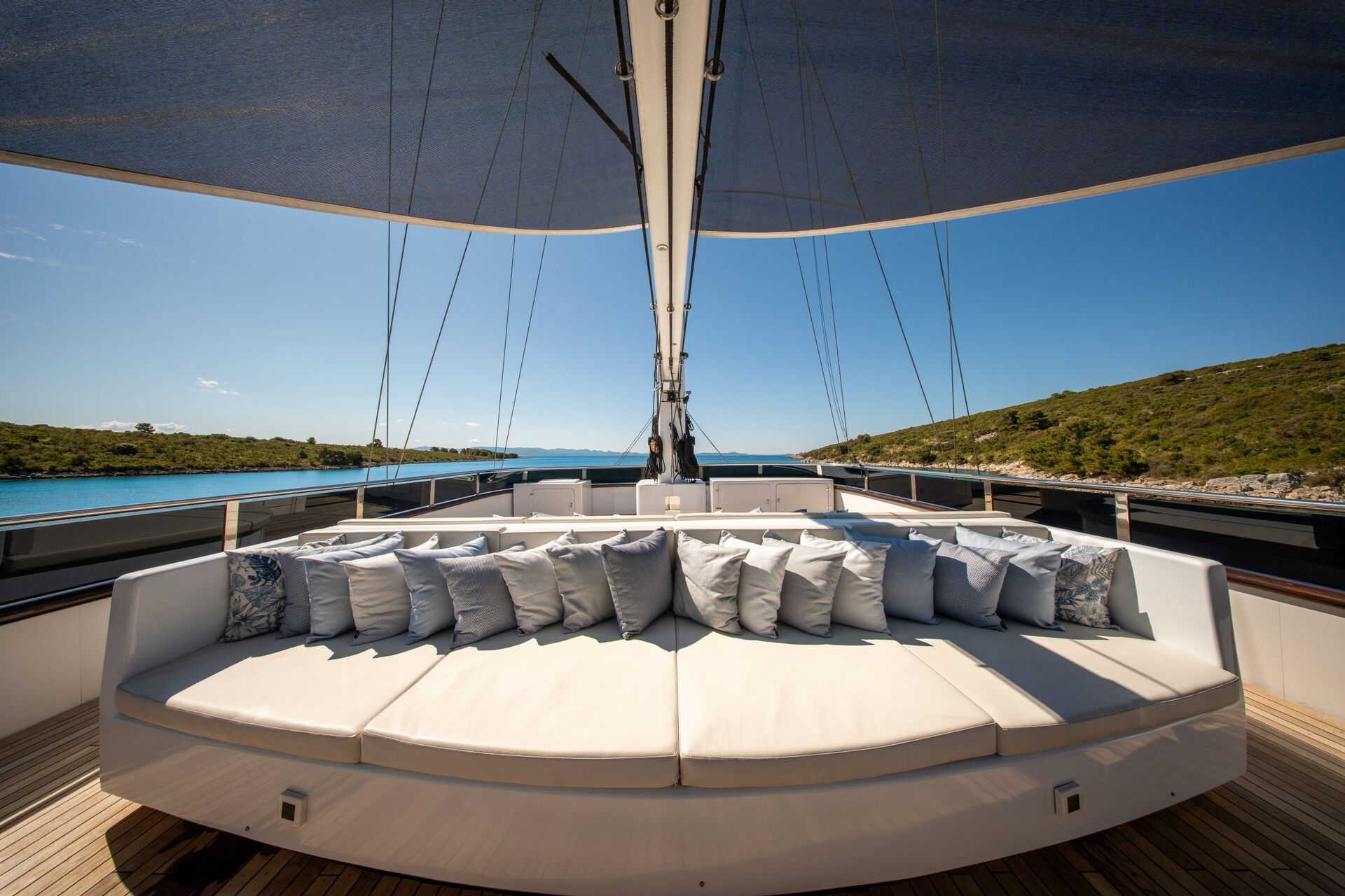 A large, circular white sofa with many pillows sits on the deck of a yacht under a canopy, with water and green hills visible on both sides under a clear blue sky.