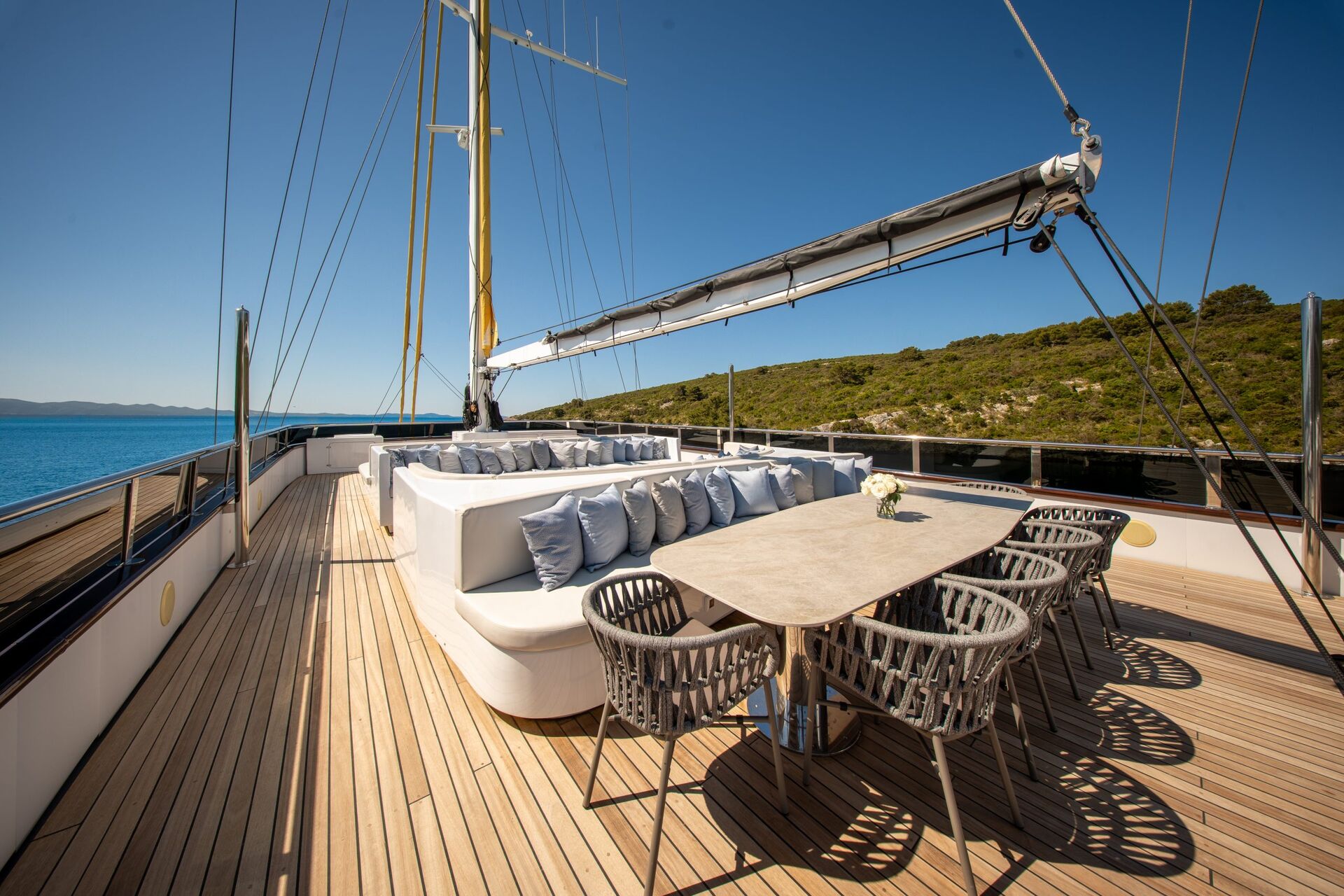 A luxurious yacht deck with cushioned seating, a dining table with six woven chairs, and a vase of flowers. The yacht is on calm blue water, near a green, hilly coastline under a clear blue sky.