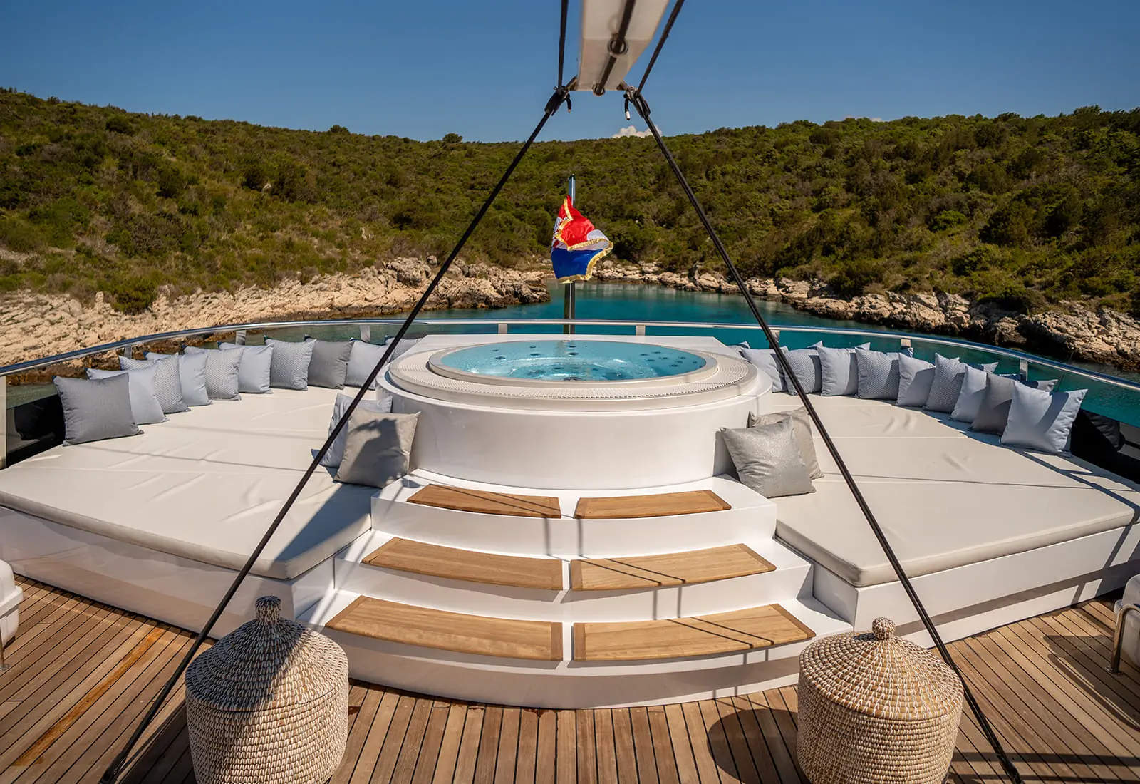 A luxury yacht deck features a circular hot tub, white cushioned seating with pillows, and wooden steps, overlooking clear blue water and green hills under a clear sky.