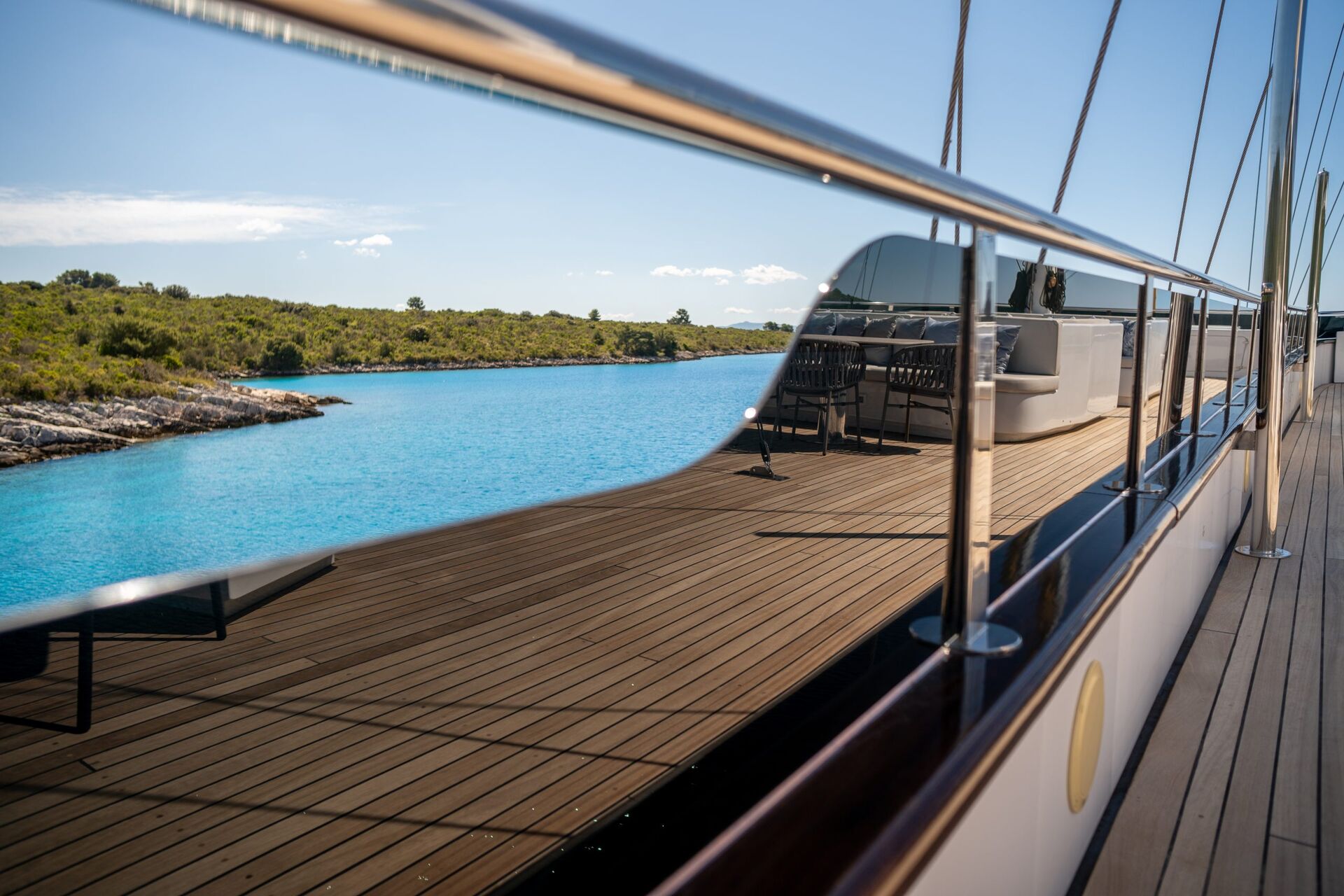 A clear blue sea and green shoreline are reflected in the shiny railing of a yacht’s wooden deck under a bright sky. Outdoor seating is visible on the yacht.