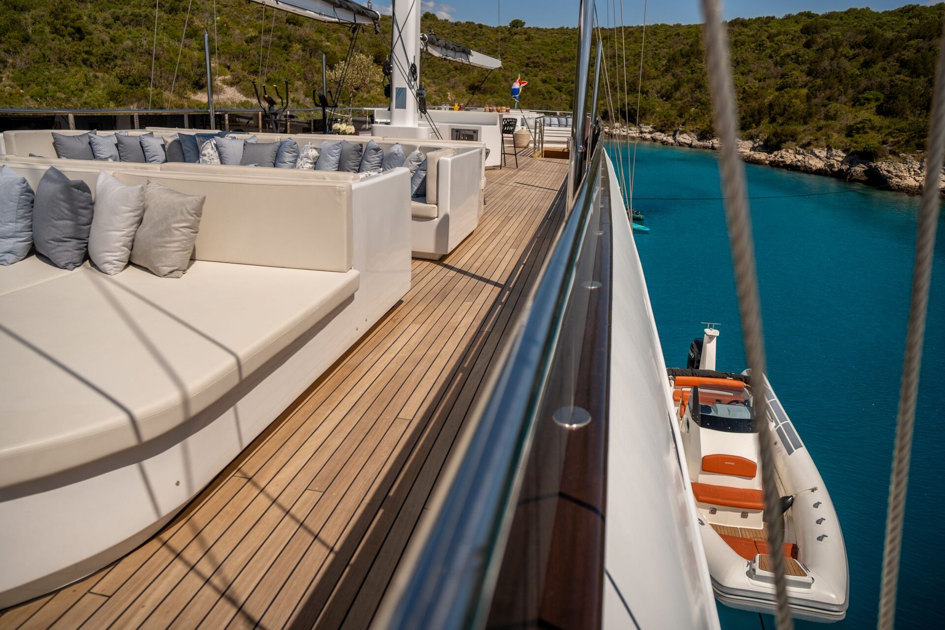 Luxury yacht deck with white cushioned seating, wooden flooring, and a view of clear turquoise water and green hills. A small boat is suspended alongside the yacht near the waters edge.