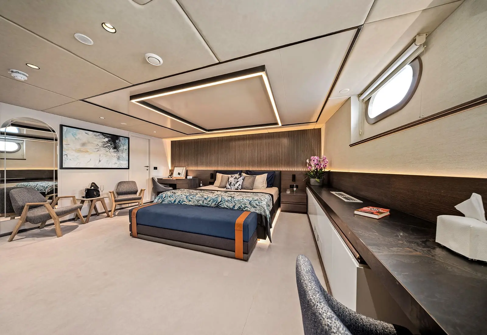 Modern bedroom with a king-size bed, wall art, two chairs, a desk, a window, and decorative lighting. There are flowers, a tissue box, and a book on the dark wood desk. The room looks spacious and stylish.