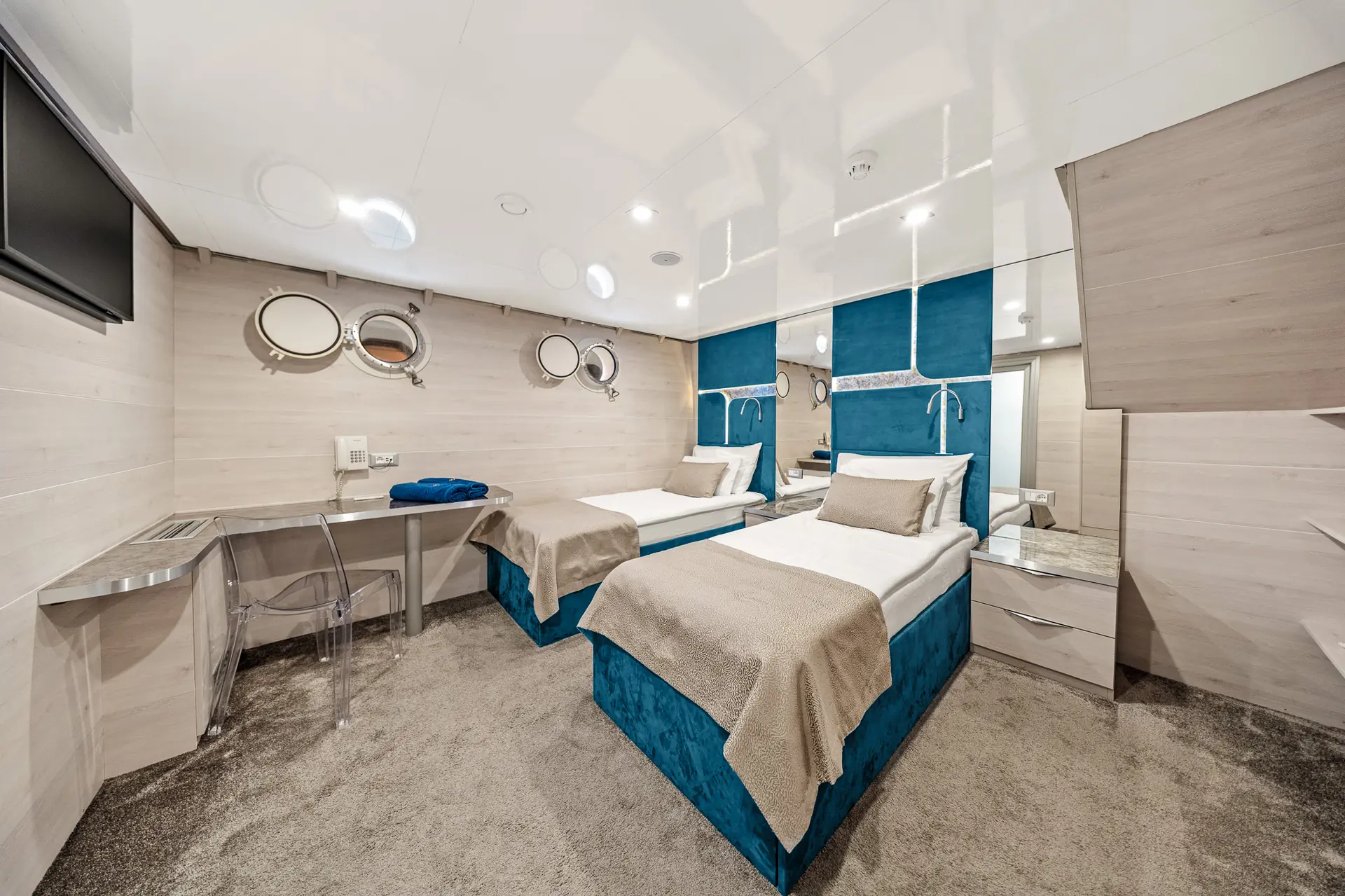 Modern cabin room with two single beds, blue headboards, round porthole windows, beige bedding, a desk with a clear chair, and mirrored walls, creating a bright and spacious atmosphere.