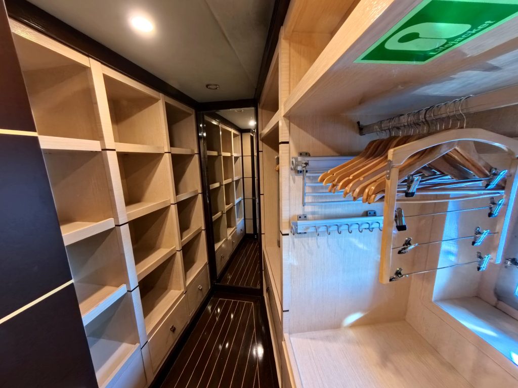 A modern walk-in closet with empty wooden shelves and hanging rods, several wooden hangers, and a sign above. The closet has a glossy dark floor and a full-length mirror at the end.