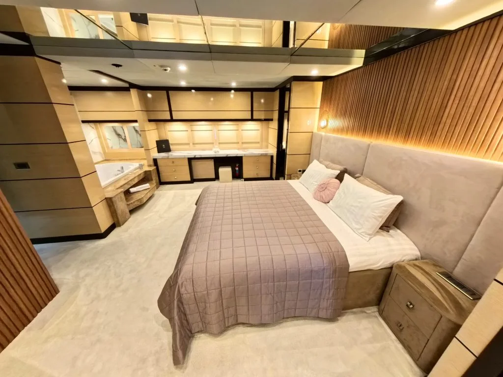 A modern bedroom with beige and wood accents features a large bed with a quilted taupe blanket, built-in bedside tables, a mirrored ceiling, and a spacious vanity area with ample cabinetry and lighting.