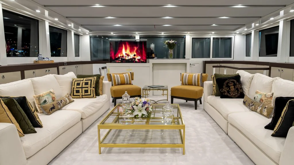 Modern yacht lounge with white sofas, gold-accented pillows, a glass coffee table with flowers, two yellow chairs, and a TV above a faux fireplace, all under bright ceiling lights. Large windows line the walls.