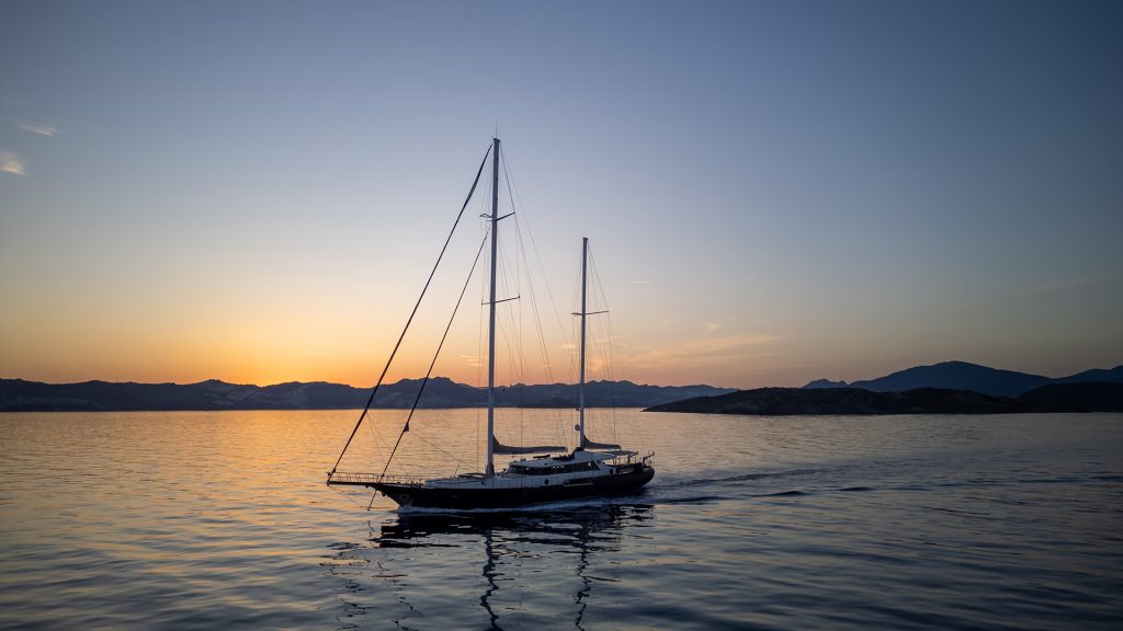 A sailboat glides on calm water at sunset, with silhouetted mountains and a colorful sky in the background.