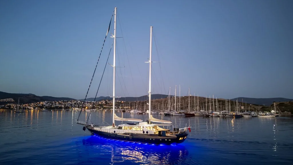 A large sailboat with illuminated blue lights underneath glides on calm water at dusk, with a marina full of docked boats and distant city lights visible in the background.