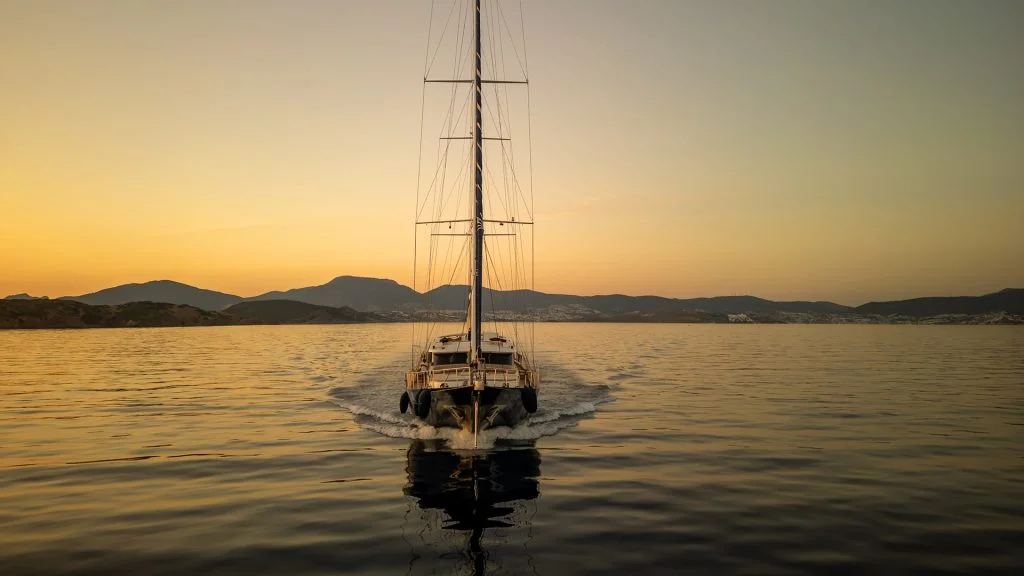 A sailboat cruises on calm water at sunset, with mountains and a distant shoreline silhouetted against the orange and yellow sky.