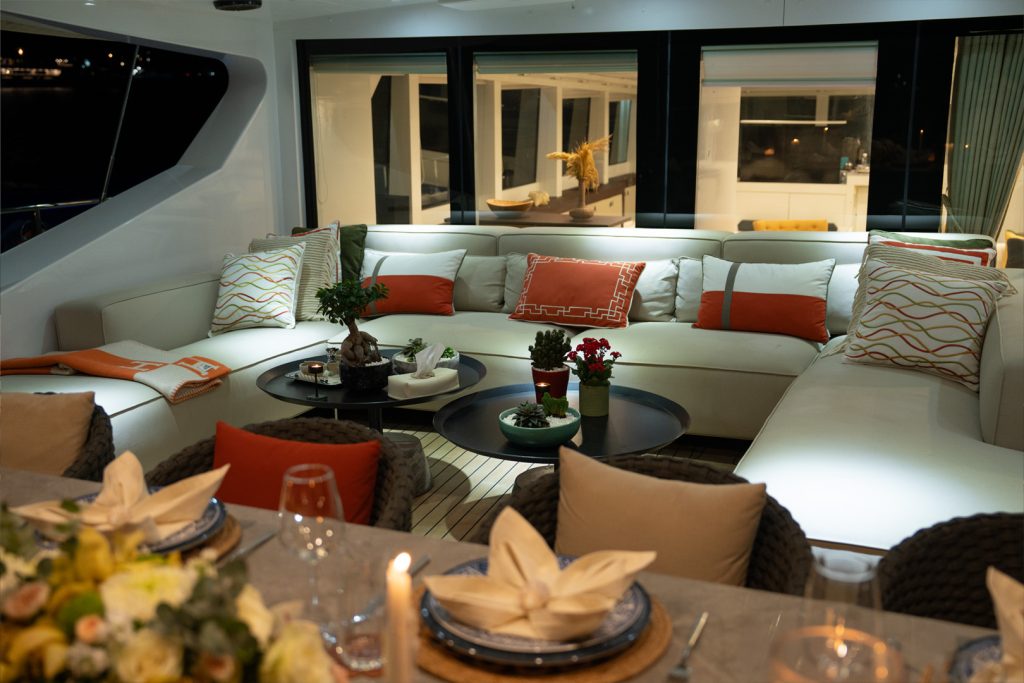 A modern yacht lounge with white sectional sofas, orange and patterned pillows, round coffee tables with plants, and a dining table set with plates, napkins, and a flower centerpiece in the foreground.