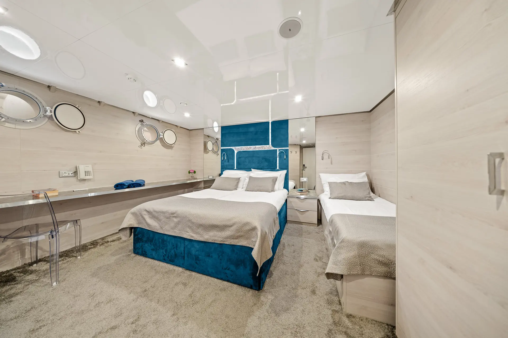 Modern ship cabin with a large double bed, a single bed, light wood-paneled walls, round porthole windows, a desk with a clear chair, and blue accents, creating a bright and cozy atmosphere.