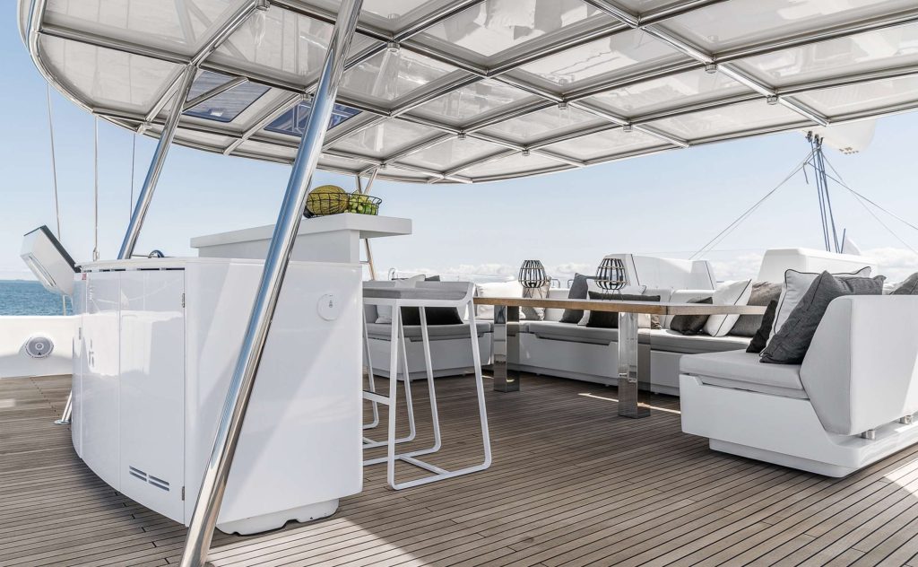 ABOVE & BEYOND Modern yacht deck with white seating, a dining table, bar area, and overhead canopy. Cushions and decor add comfort. Ocean and blue sky visible in the background.