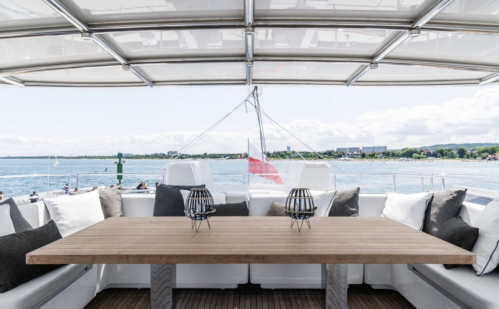 ABOVE & BEYOND A modern yacht deck with a wooden table, decorative lanterns, and cushioned seating, overlooking calm water and a distant shoreline under a partly cloudy sky. A flag is visible at the back of the yacht.